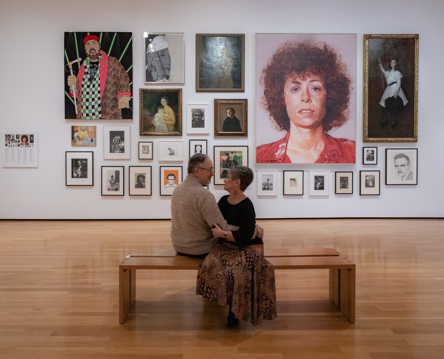Gallery visitors view a wall display of framed photographs in a museum exhibition space with wooden benches.