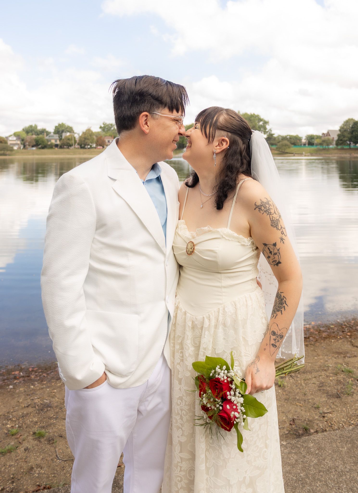 Wedding couple poses by lakeside with red bouquet against cloudy sky backdrop.