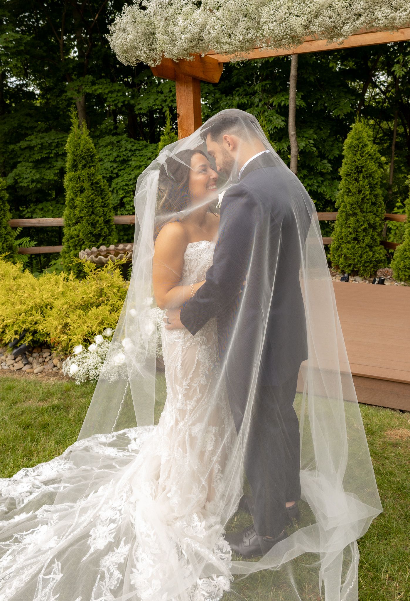 Romantic wedding couple embracing under flowing white veil in garden setting.