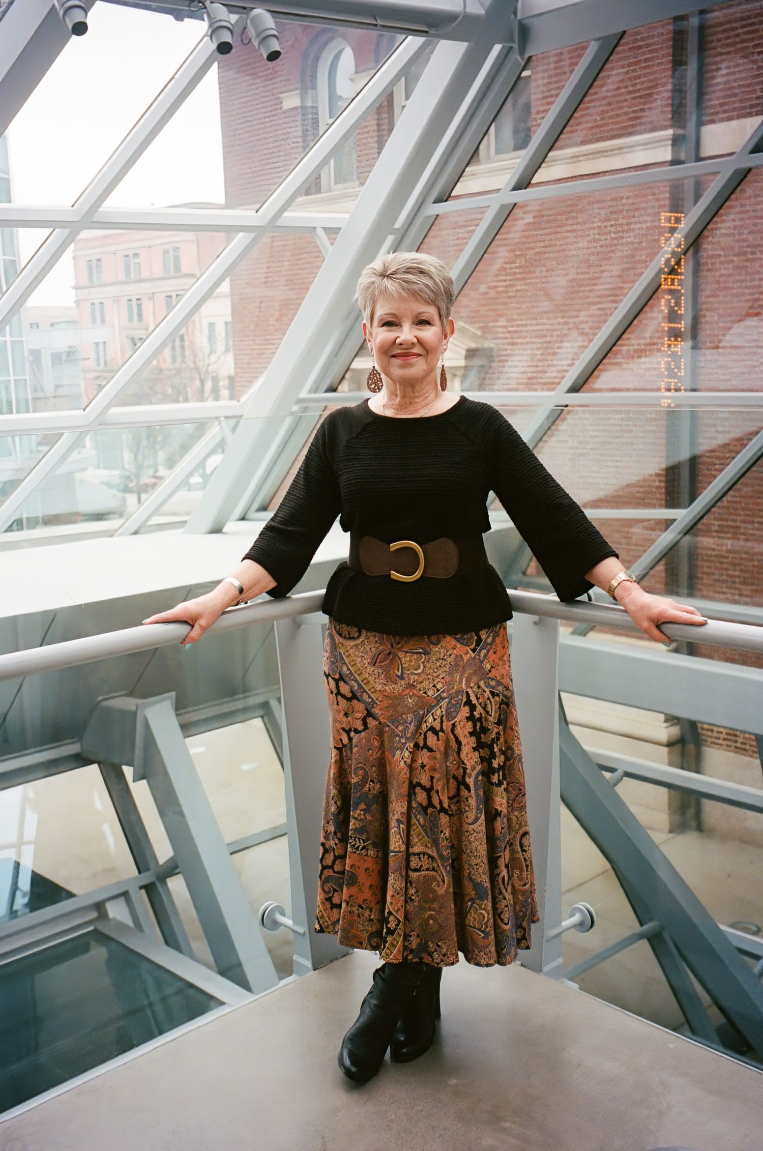 A person in a black top and patterned skirt poses in a modern architectural space with angled glass windows.