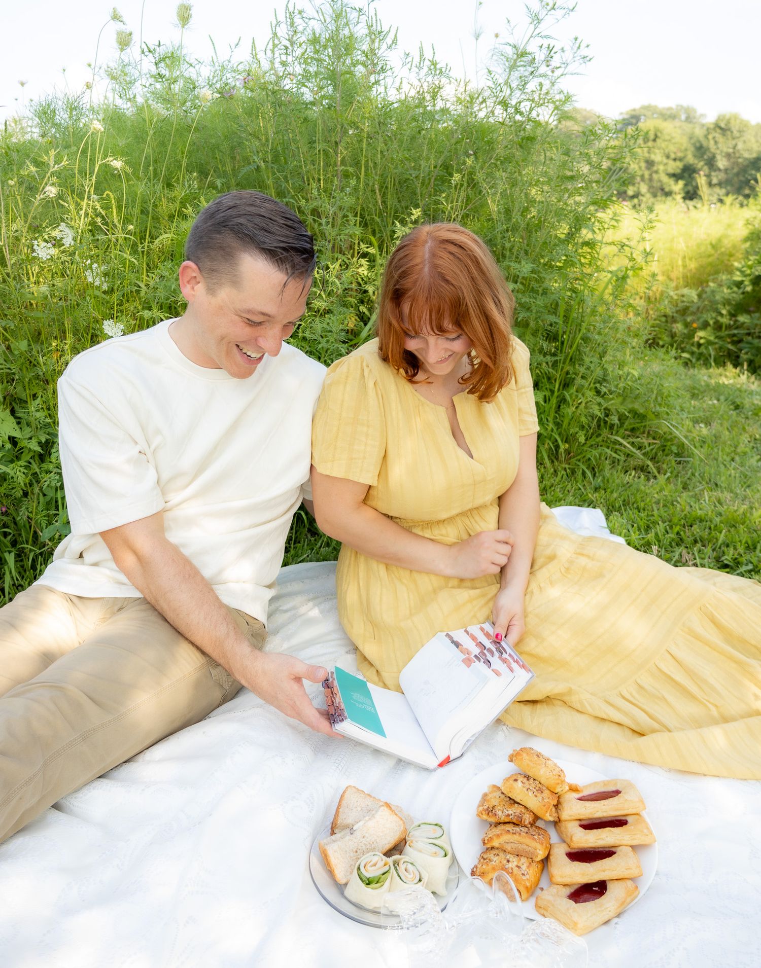 A couple enjoys a romantic outdoor picnic with sandwiches and drinks on a white blanket in a grassy field.
