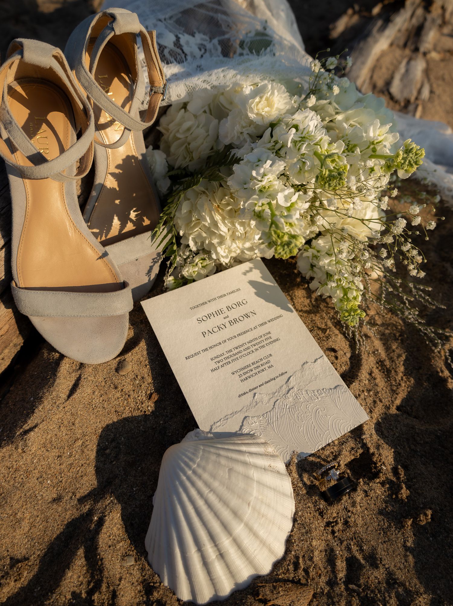 Wedding invitation cards displayed on sand with seashells and white flowers as decoration.