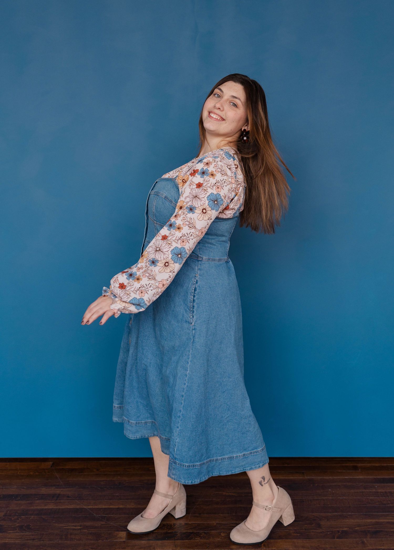 Person in denim dress and floral top posing playfully against blue backdrop.