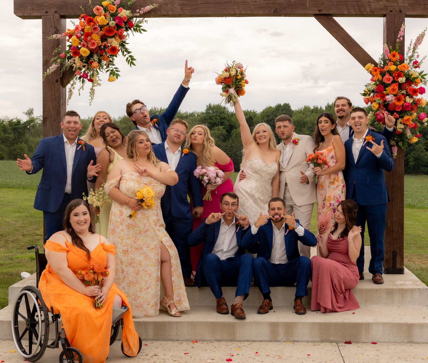Large wedding party posing together under wooden arch decorated with orange and red fall flowers.