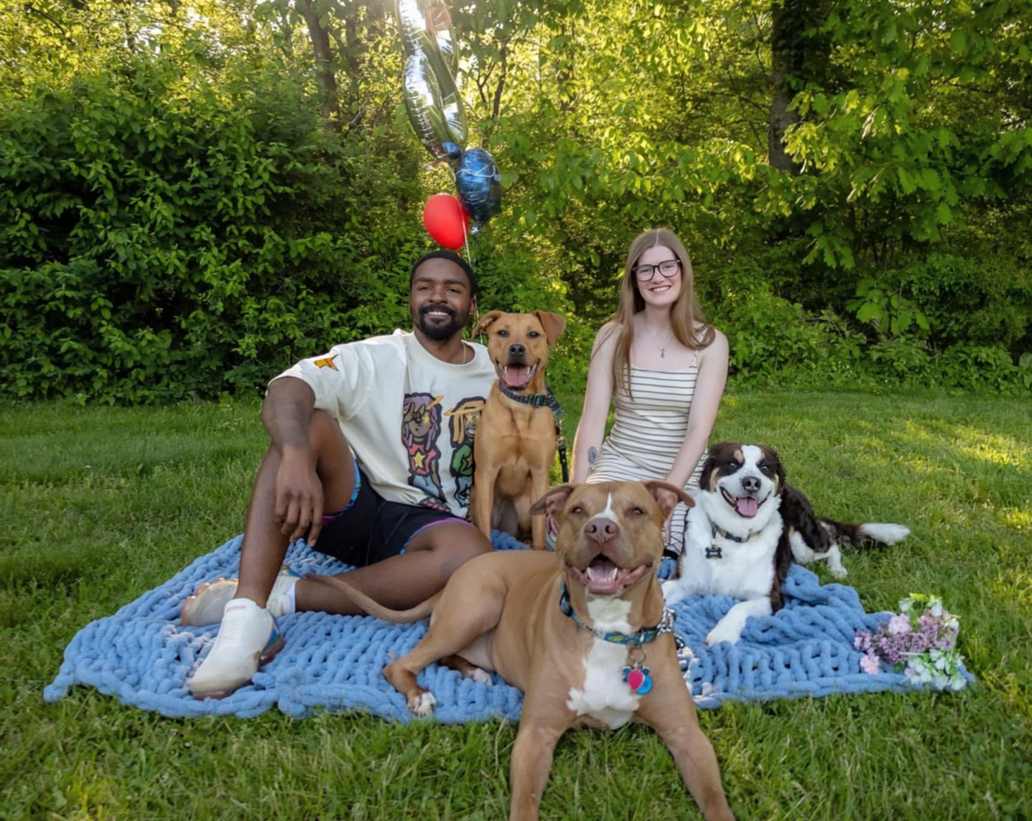 Group enjoying a summer picnic on a blue blanket with their dog in a lush green park setting.