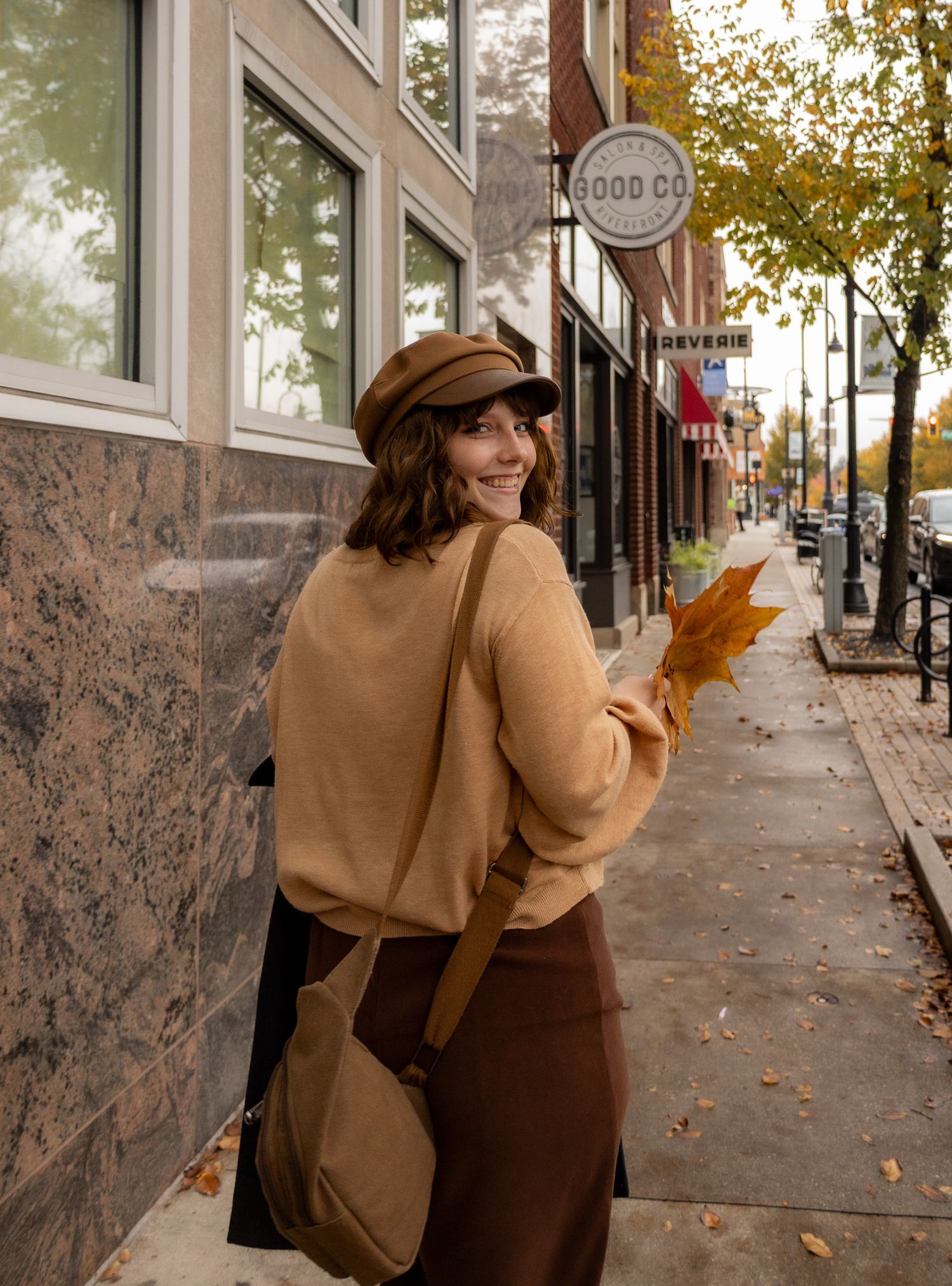 Person in tan sweater walking down an autumn city sidewalk with fallen leaves.
