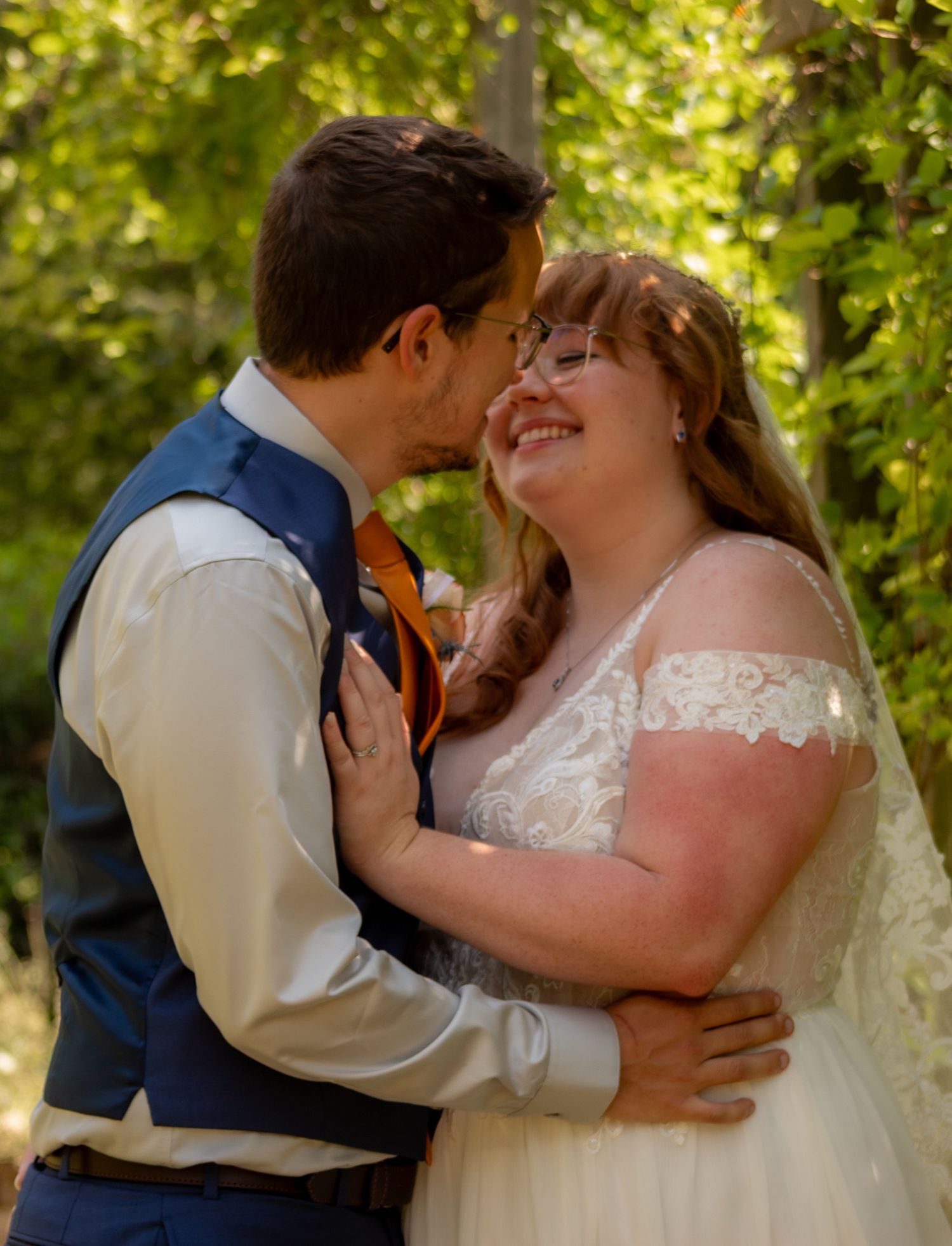 Romantic wedding couple embracing outdoors surrounded by green summer foliage.