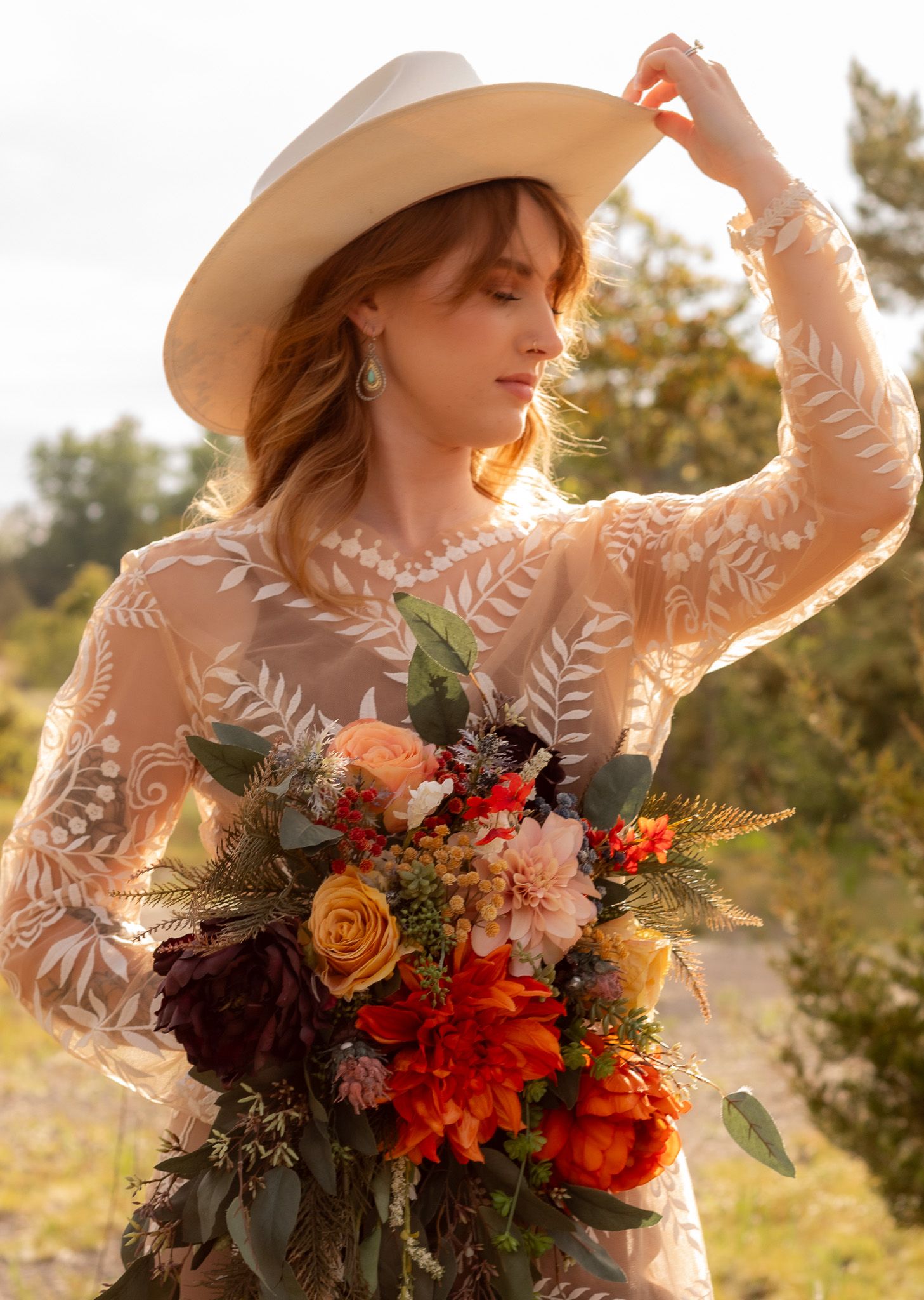 Ethereal autumn bride in lace dress and wide brim hat holds vibrant orange and burgundy floral bouquet at sunset.