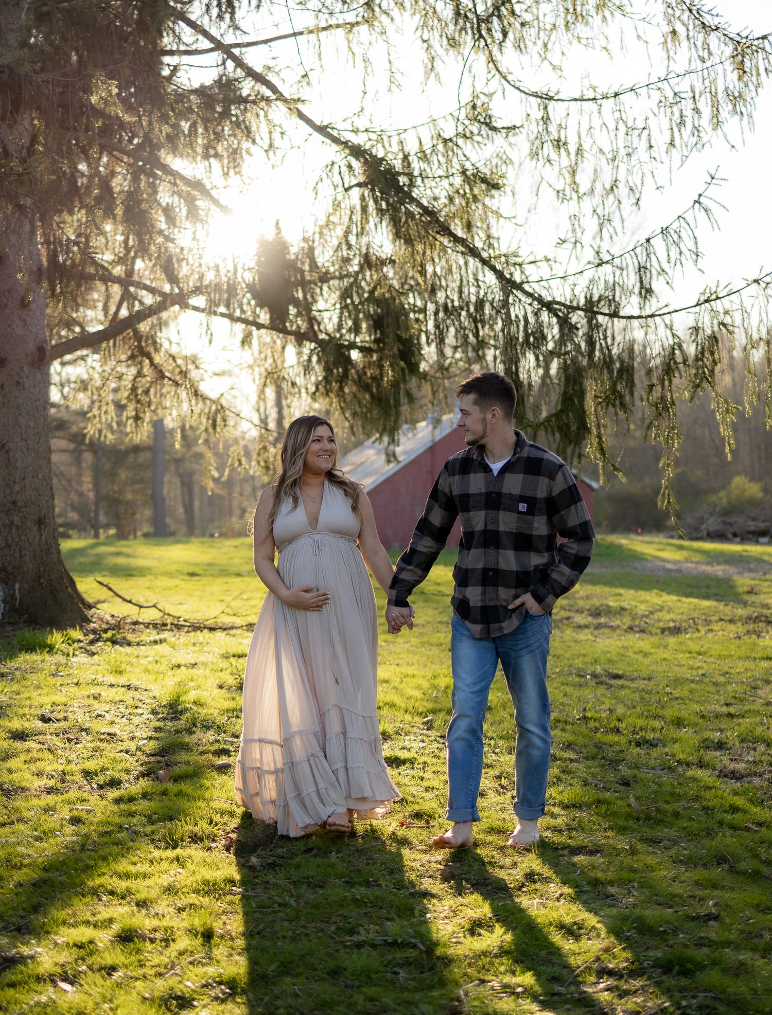 Couple in a dreamy outdoor maternity photoshoot with sunlit evergreen trees and lush grass in the background.