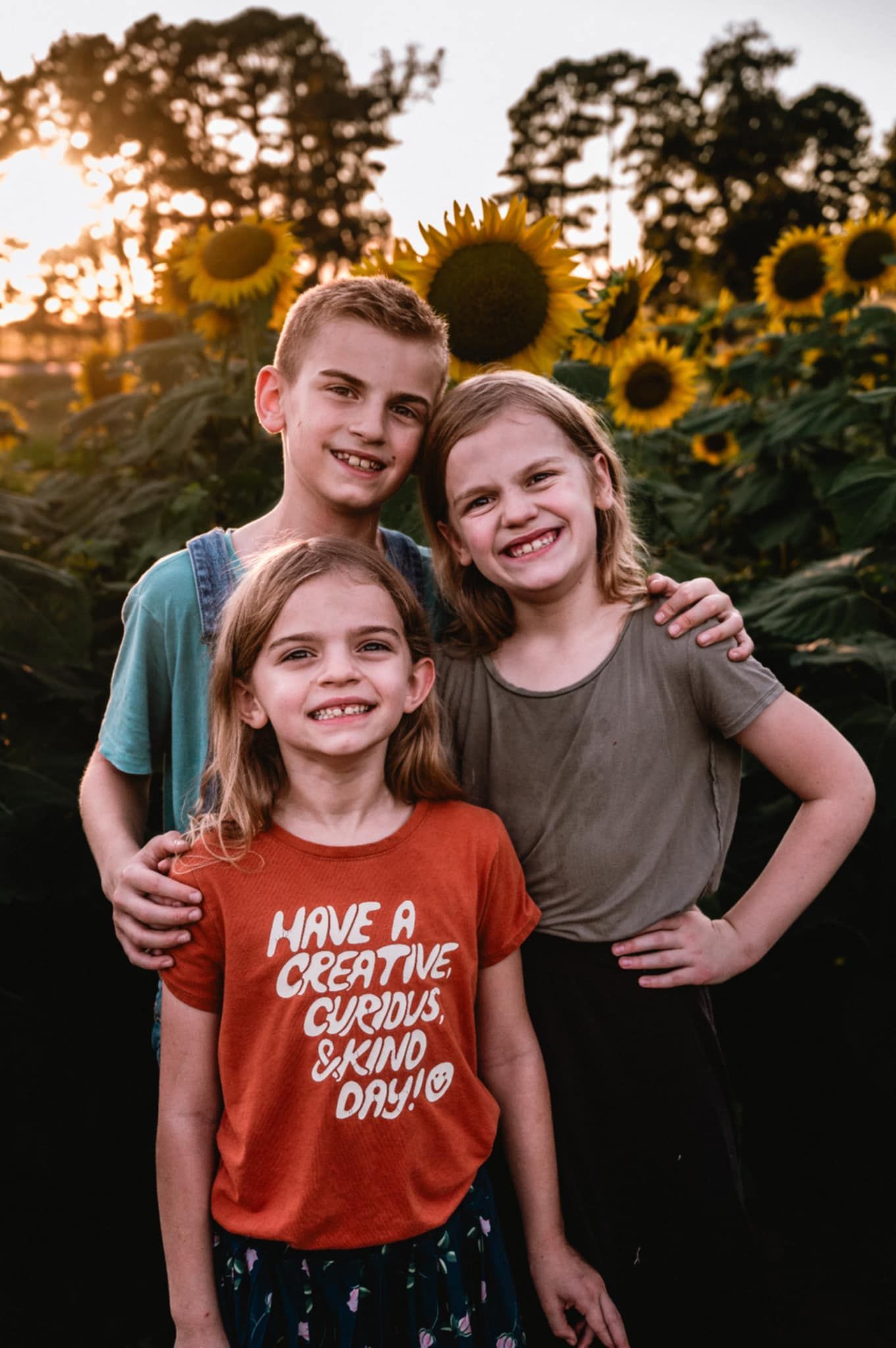 Three children pose together in front of blooming sunflowers at sunset with golden light filtering through trees.