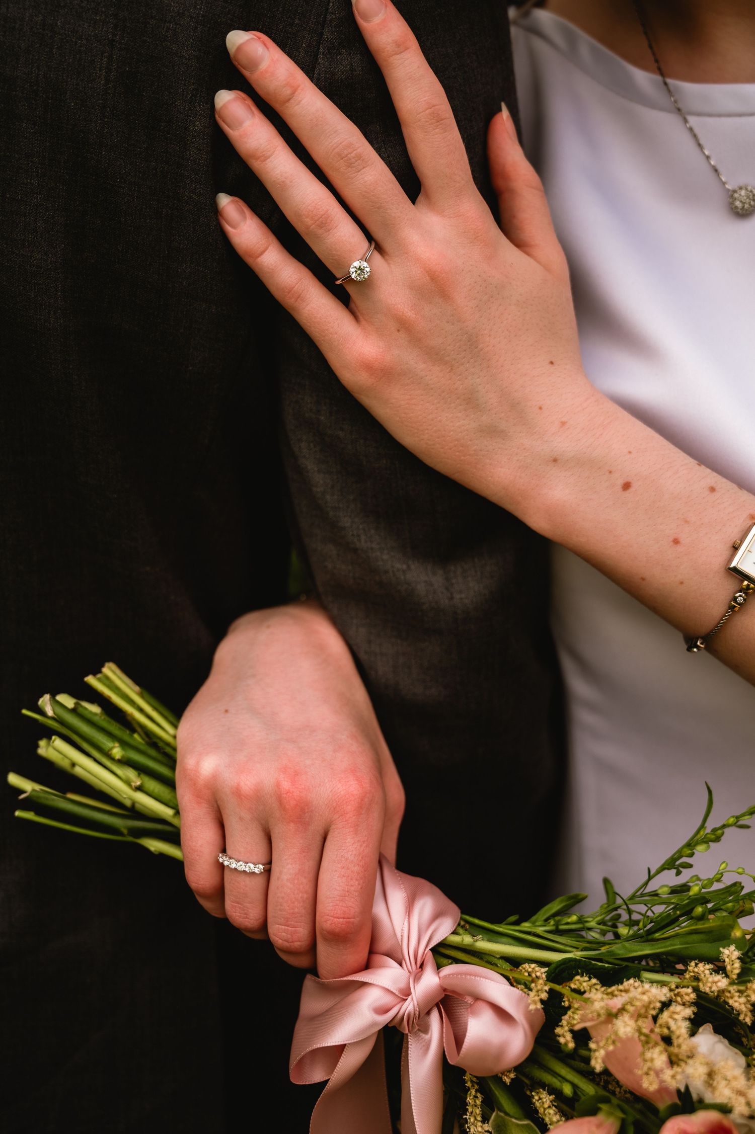 Bride showing engagement ring on hand resting on groom's arm while holding floral bouquet with pink ribbon.