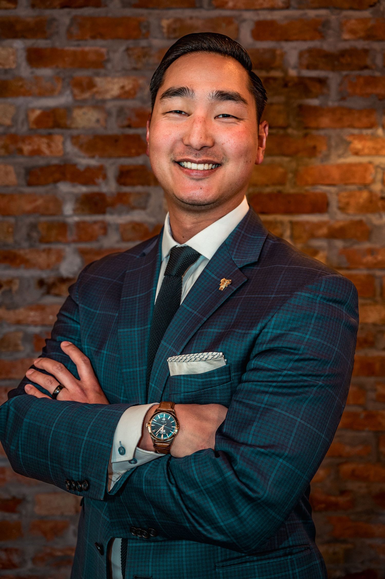 Smiling Asian man in plaid teal suit with arms crossed, posing confidently against a rustic brick wall backdrop.