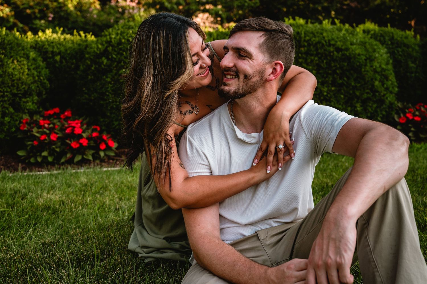 Couple sitting on green grass in a garden, woman hugging man from behind near red flowers.
