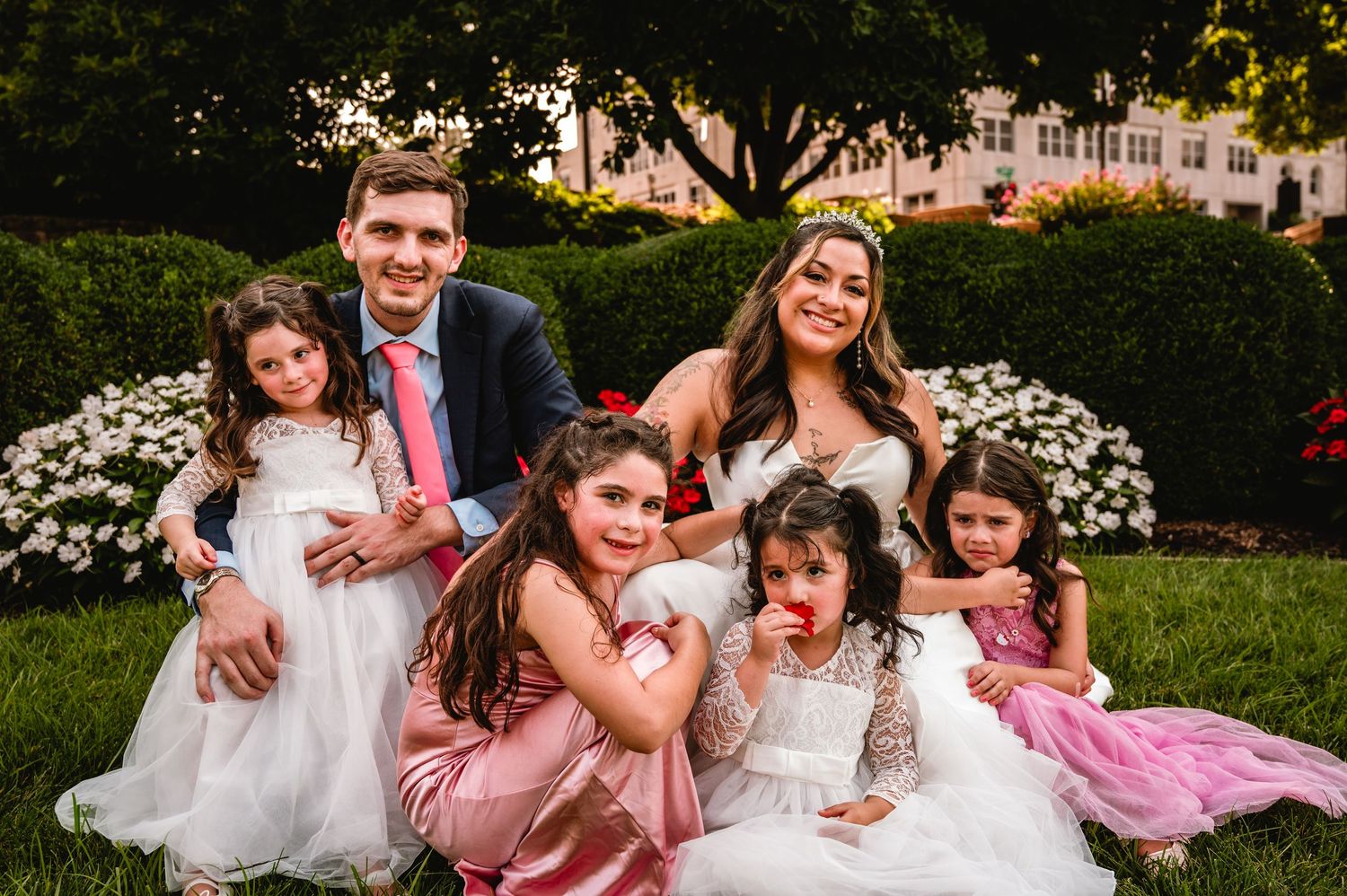 Bride and groom pose with flower girls in white dresses on green lawn with blooming flowers.