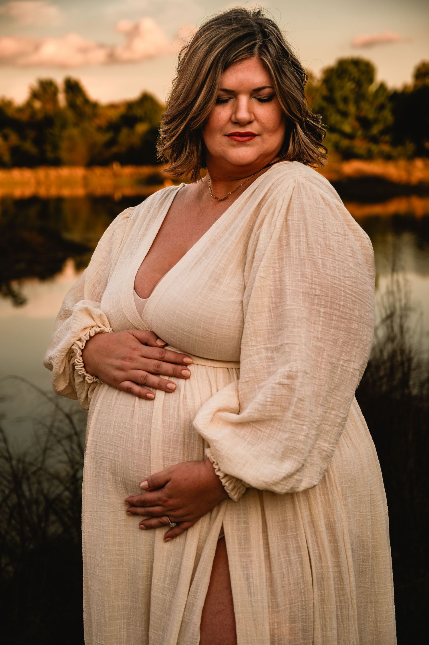 Maternity photo at sunset by a lake featuring a flowing cream-colored dress with billowy sleeves in warm golden light.