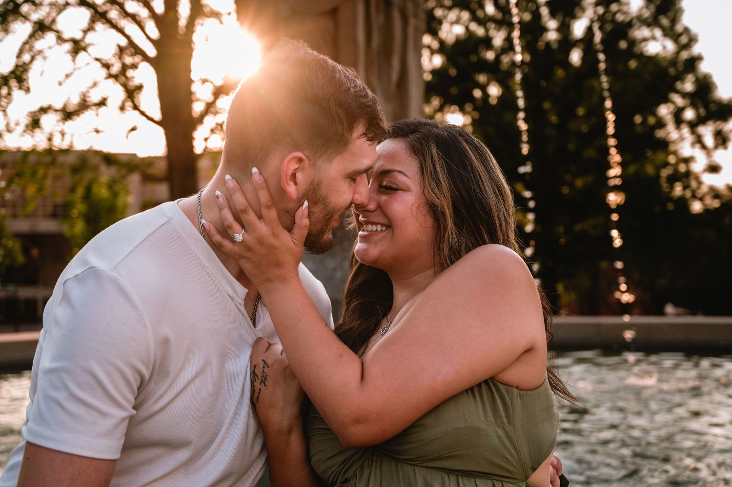 Couple sharing a romantic moment at sunset, woman smiling joyfully as man kisses her cheek near a fountain.