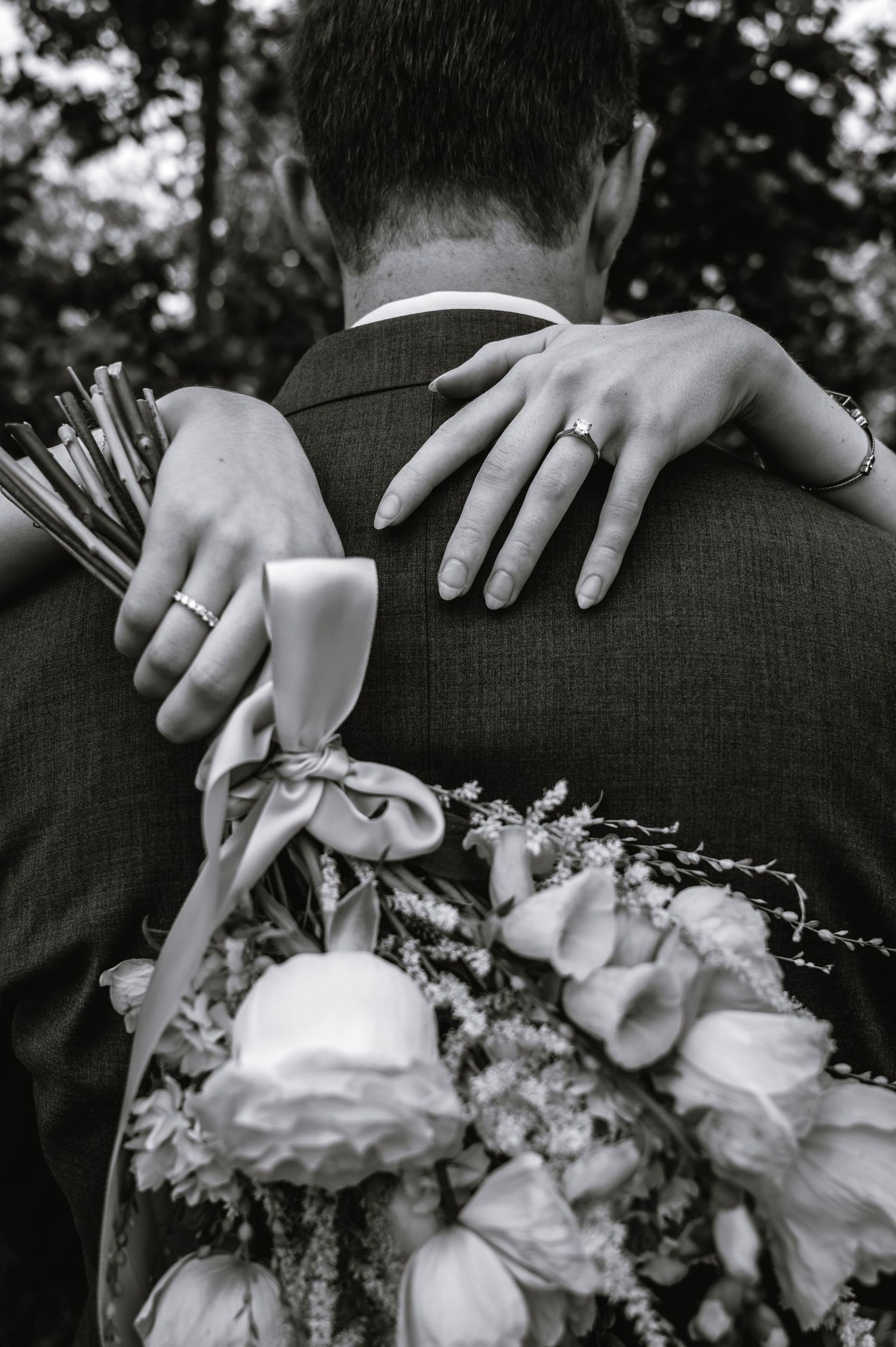 Black and white wedding photo of bride holding bouquet, arms wrapped around groom showing rings.