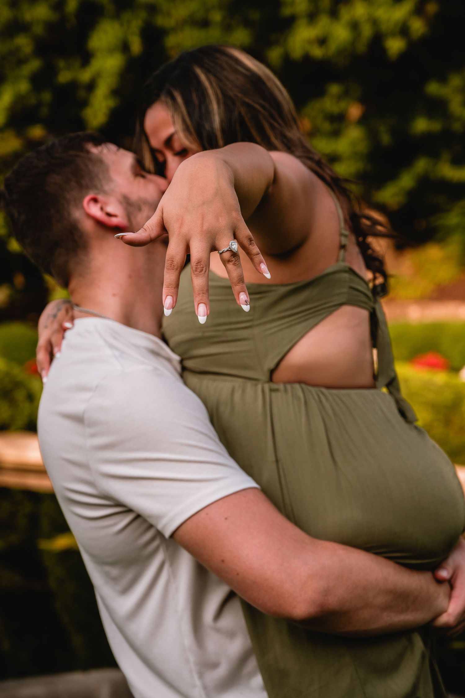 Couple embracing outdoors, woman in green dress showing engagement ring, romantic proposal moment in garden setting.