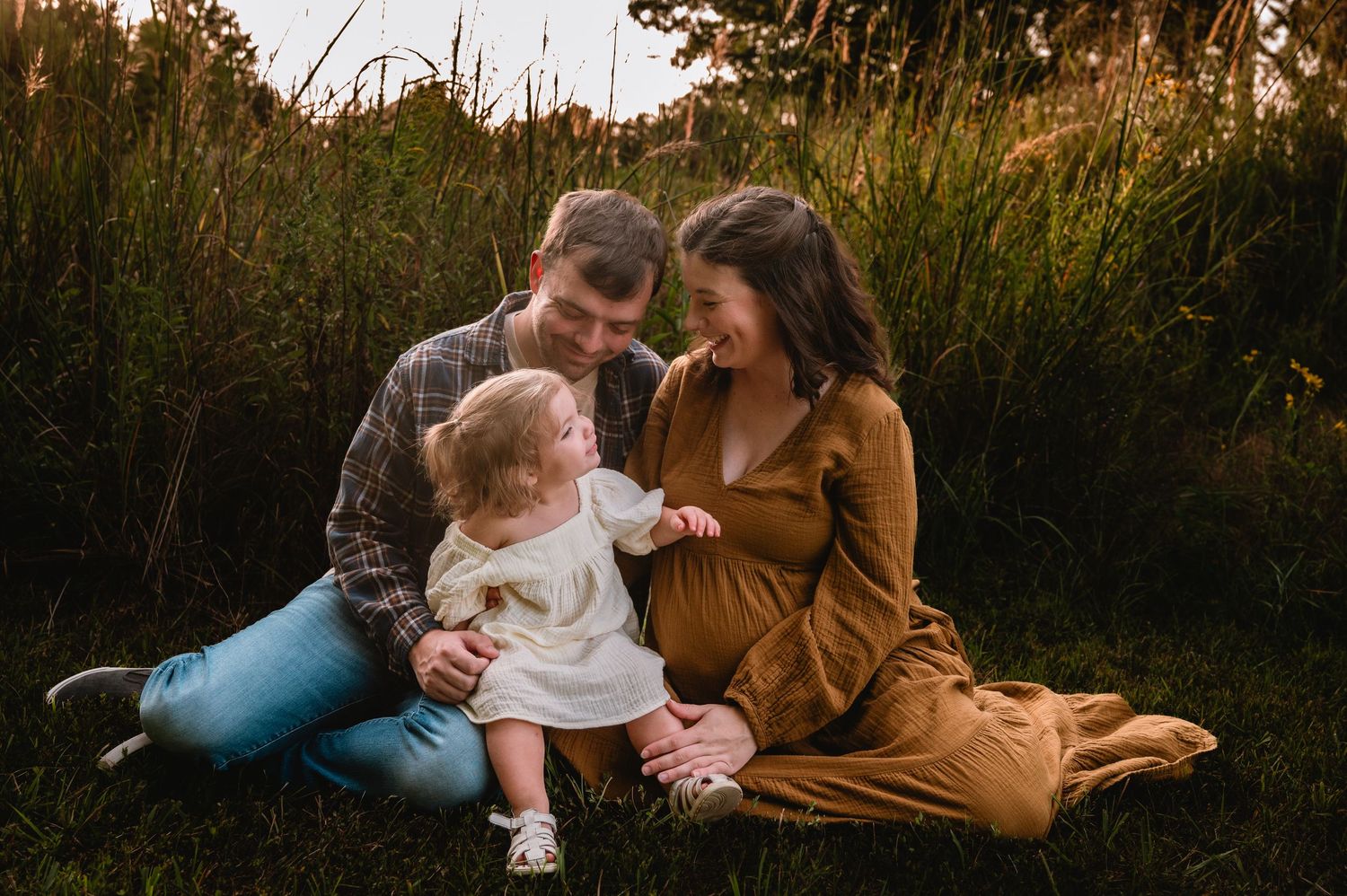 Expecting family sits in golden hour field, parents smiling at toddler daughter, maternity photo session outdoors.