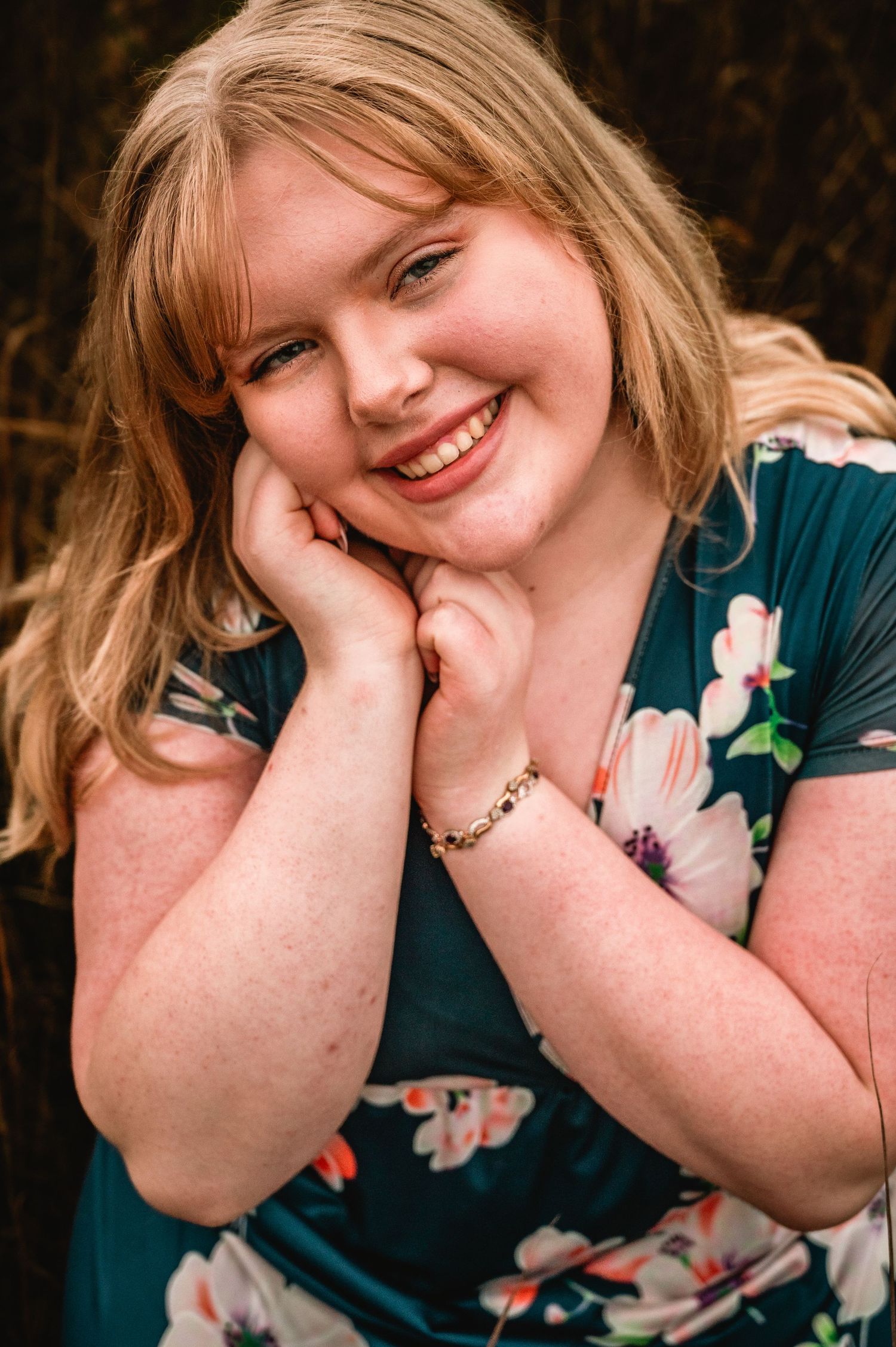 Portrait photo of someone wearing a teal floral dress while resting their head on their hand and smiling warmly.
