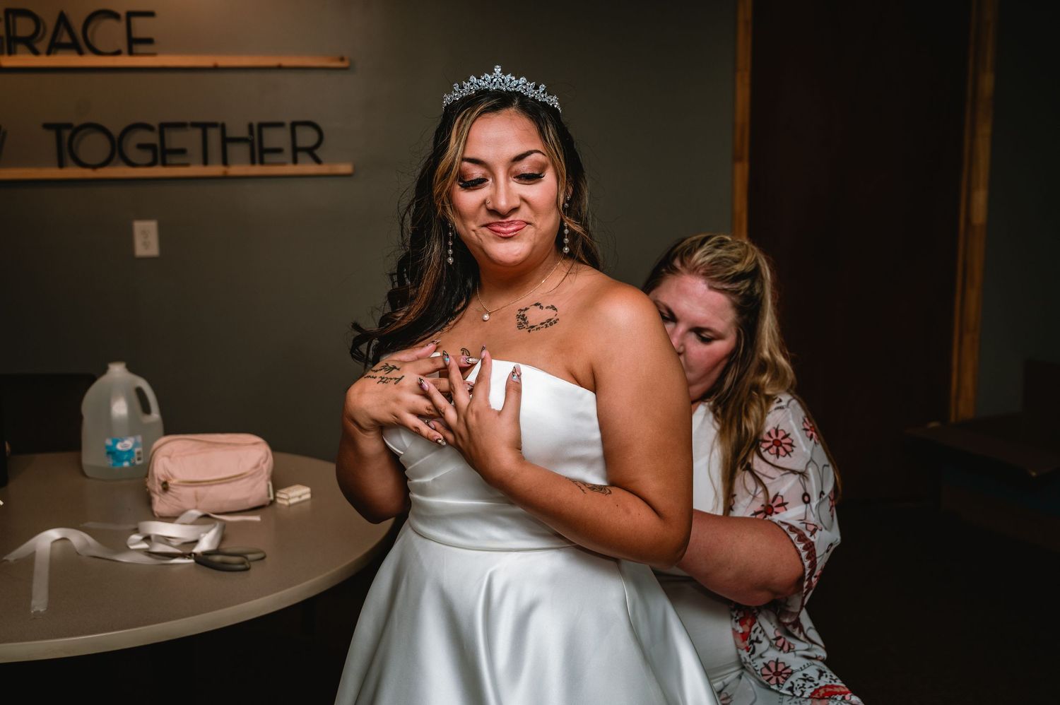 Bride in white gown and tiara gets help with necklace from bridesmaid before wedding ceremony.