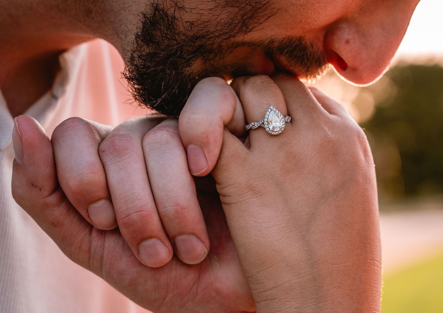 Man kissing woman's hand wearing a pear-shaped diamond engagement ring in a romantic outdoor setting.
