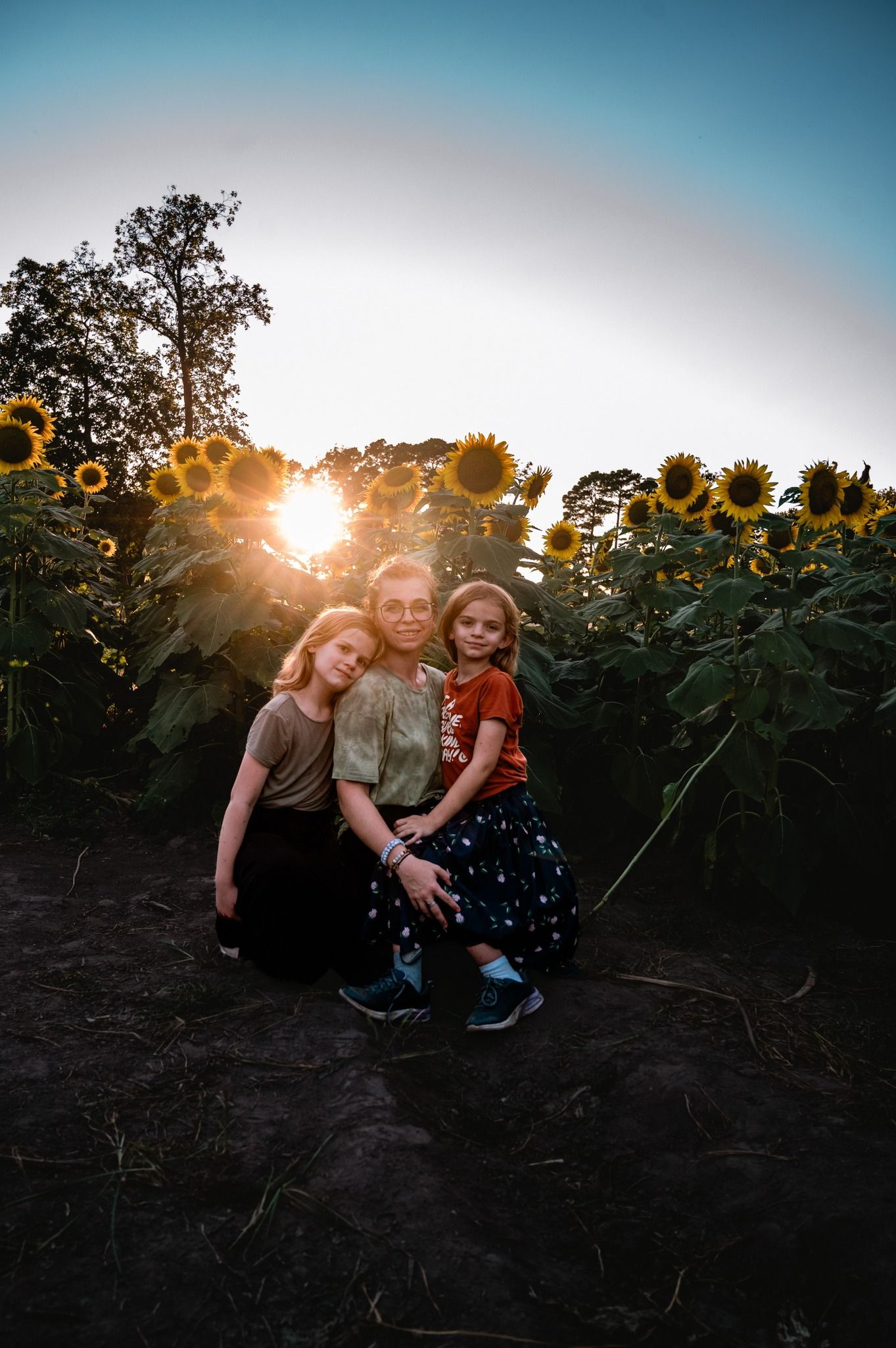 Three people sit together in a sunflower field at sunset creating a silhouette against the golden sky.