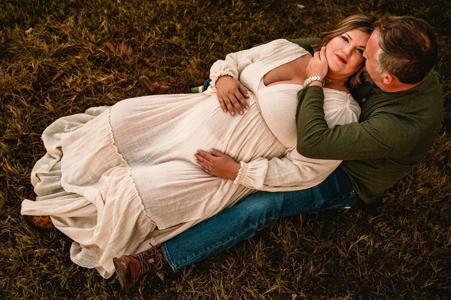 A couple in casual attire lies together on dark grass, creating an intimate outdoor portrait series.