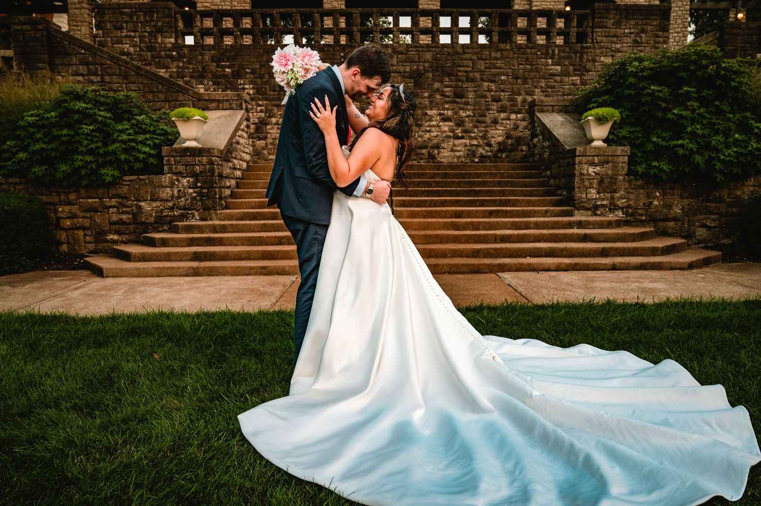 Bride and groom sharing a romantic kiss outdoors, with a flowing white wedding gown train on lush green grass.