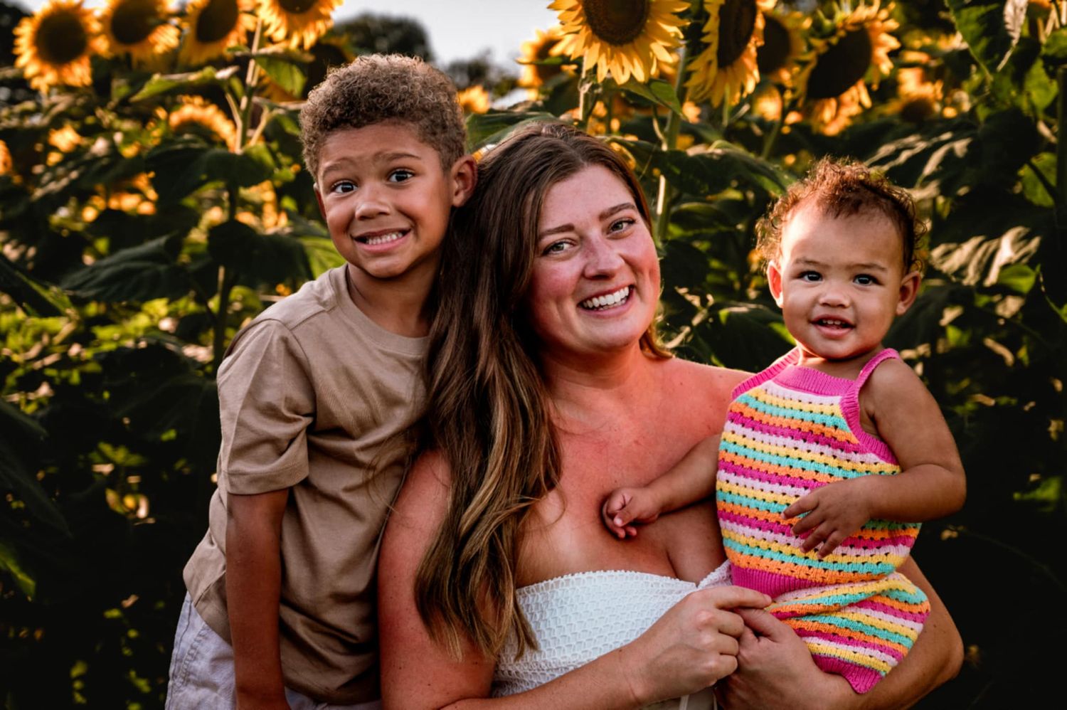 Happy family poses together in front of vibrant yellow sunflower field during golden hour.