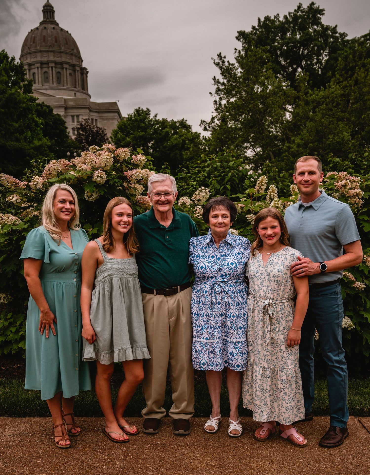 Family of six posing outdoors near a government building with blooming white hydrangeas in the background.