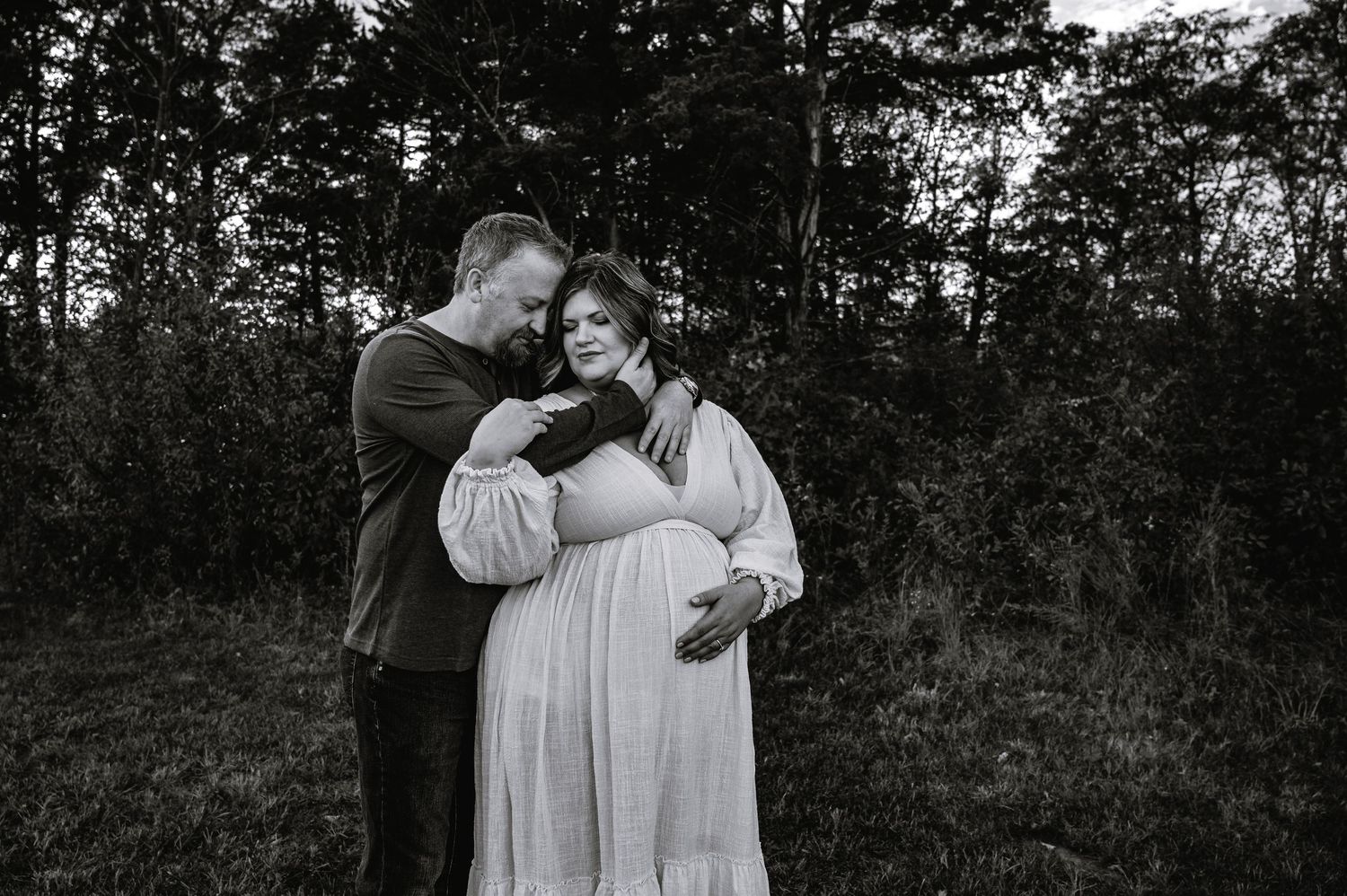 A romantic couple embraces in a wooded outdoor setting during a photo session at sunset.