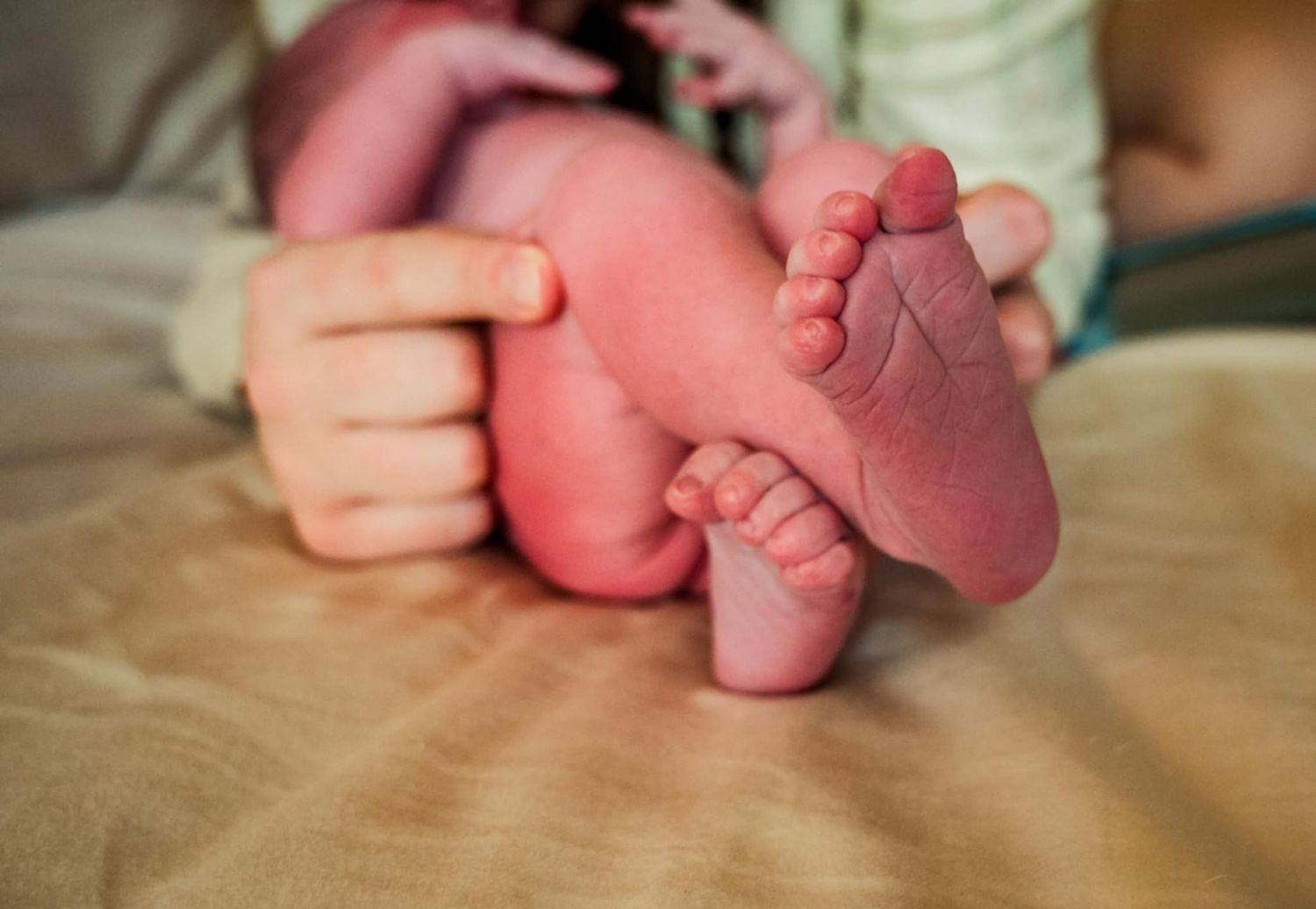 Small hands gently hold a newborn baby's tiny pink feet on a beige blanket.