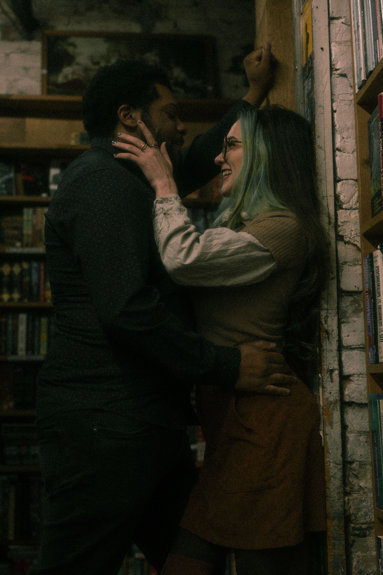 A couple embraces in a romantic dance pose against a rustic brick wall in moody lighting.