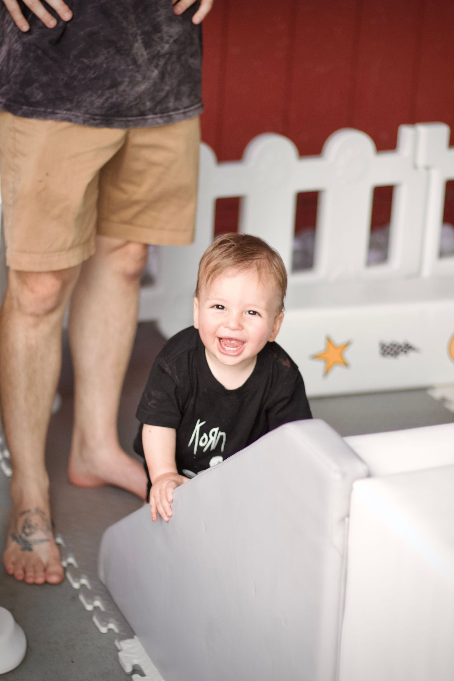 A joyful toddler in a black t-shirt plays on a white piece of furniture next to a decorative picket fence.