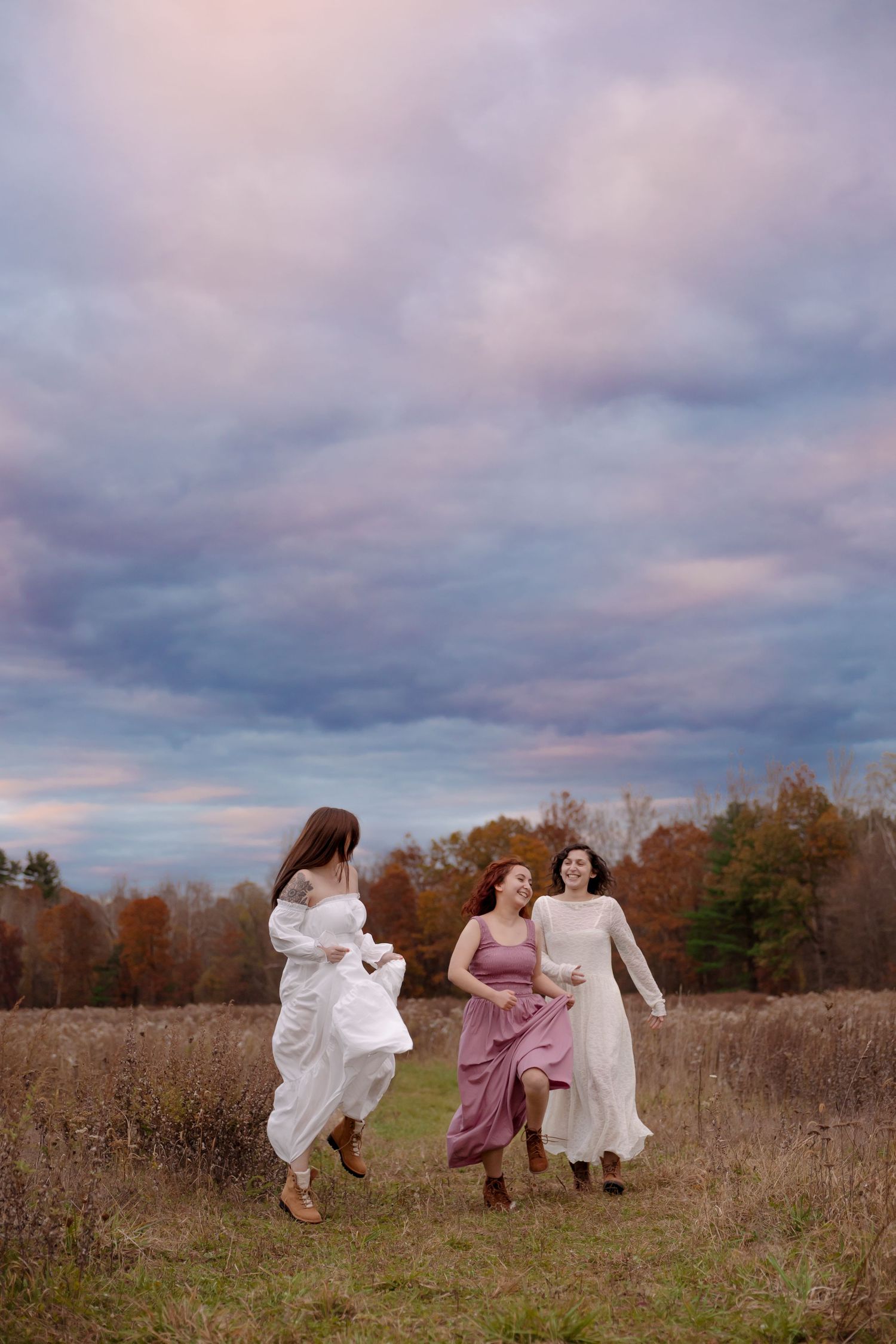 People in flowing dresses run and dance through a rustic autumn field under moody cloudy skies at sunset.