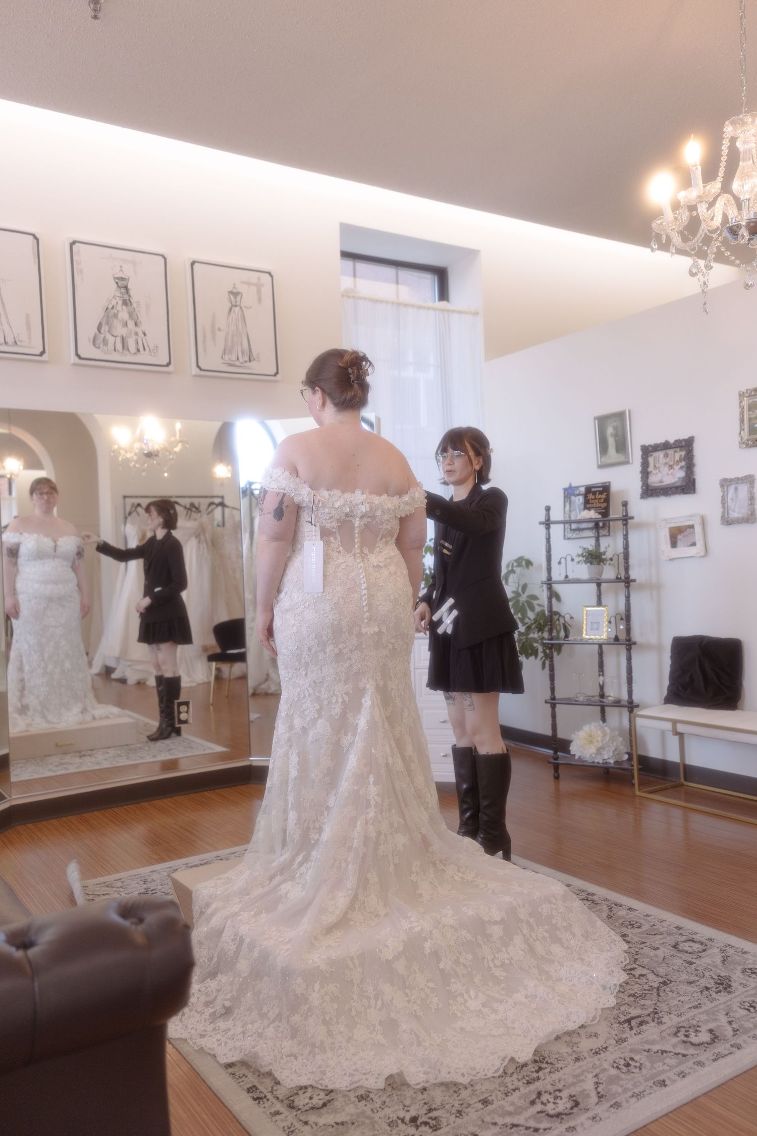 Bridal consultant adjusting a lace off-shoulder wedding gown on a bride in an elegant bridal boutique.