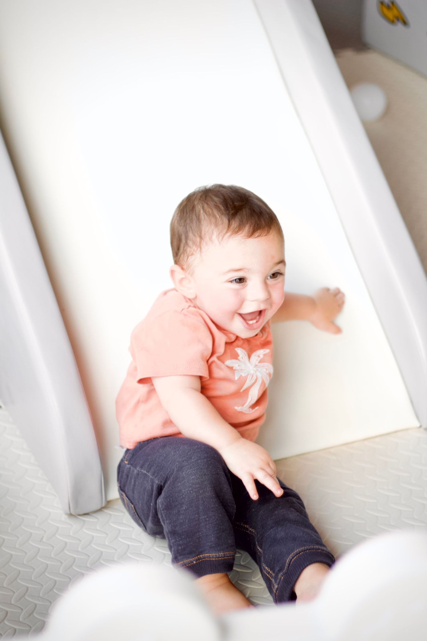 A baby wearing a coral shirt and dark pants laughs while sitting on white carpet next to a baseboard.