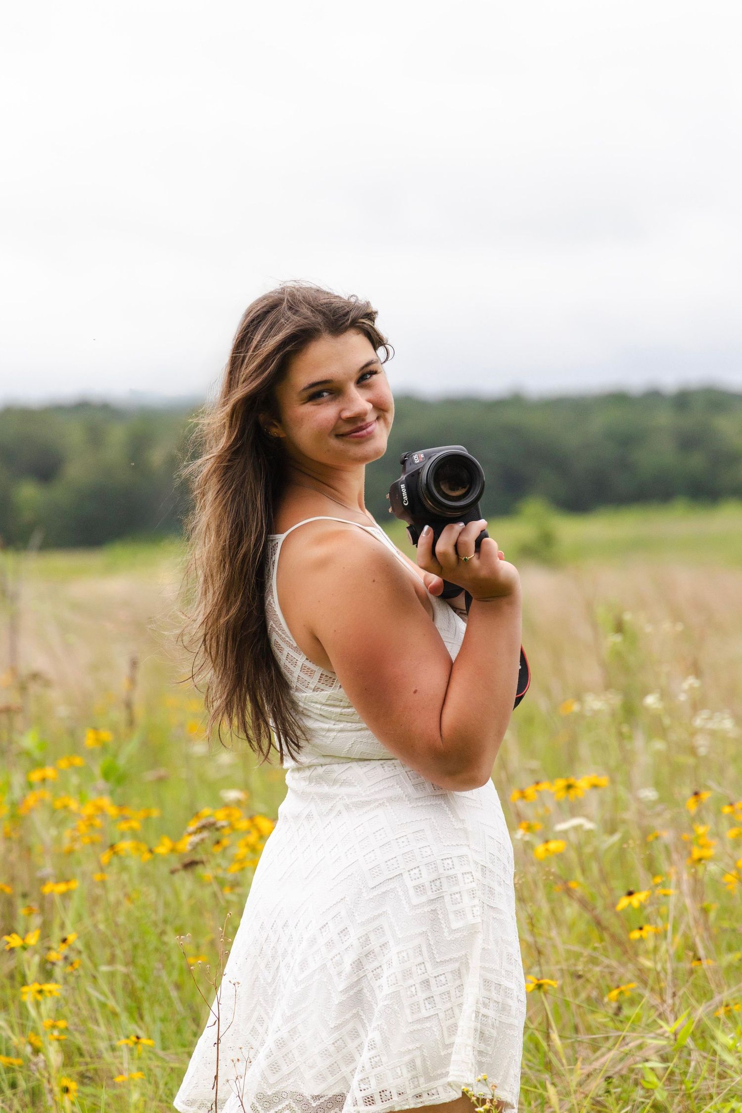 Photographer in a white lace dress holds a camera while standing in a summer wildflower meadow.