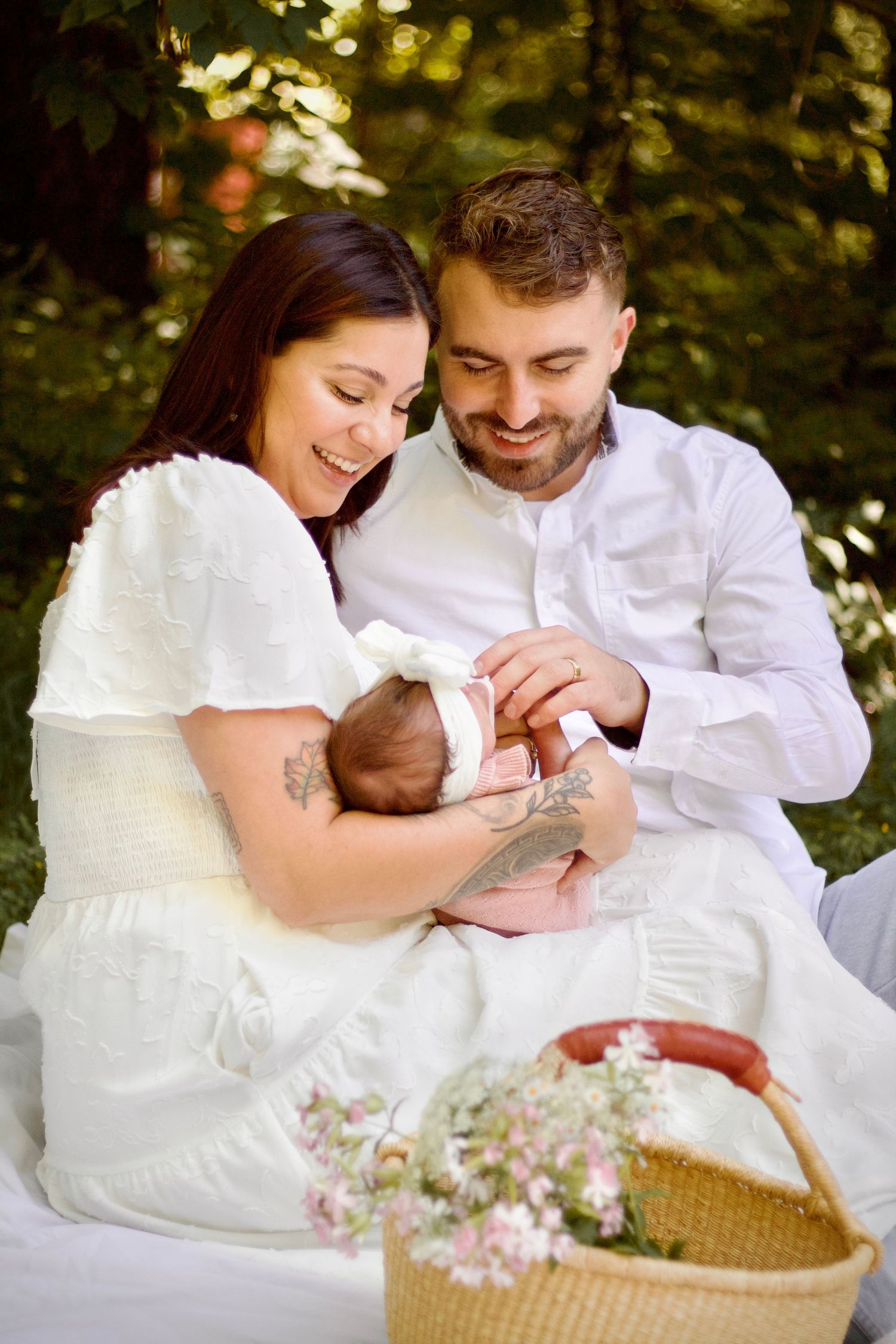 A couple in white clothing sits outdoors cradling a newborn baby next to a wicker basket with pink flowers.