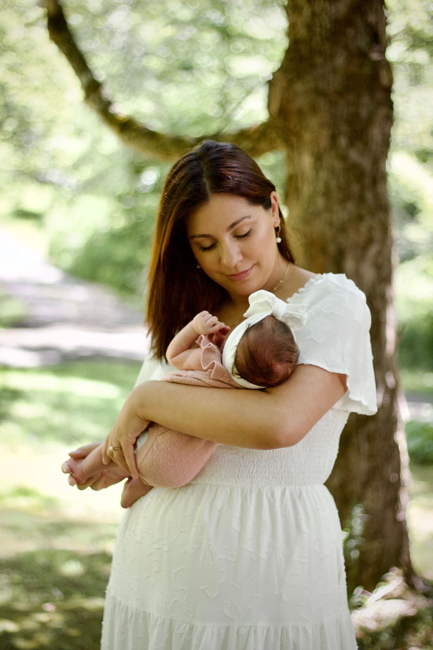 Mother in white dress holds sleeping newborn baby in outdoor garden setting with soft natural lighting.
