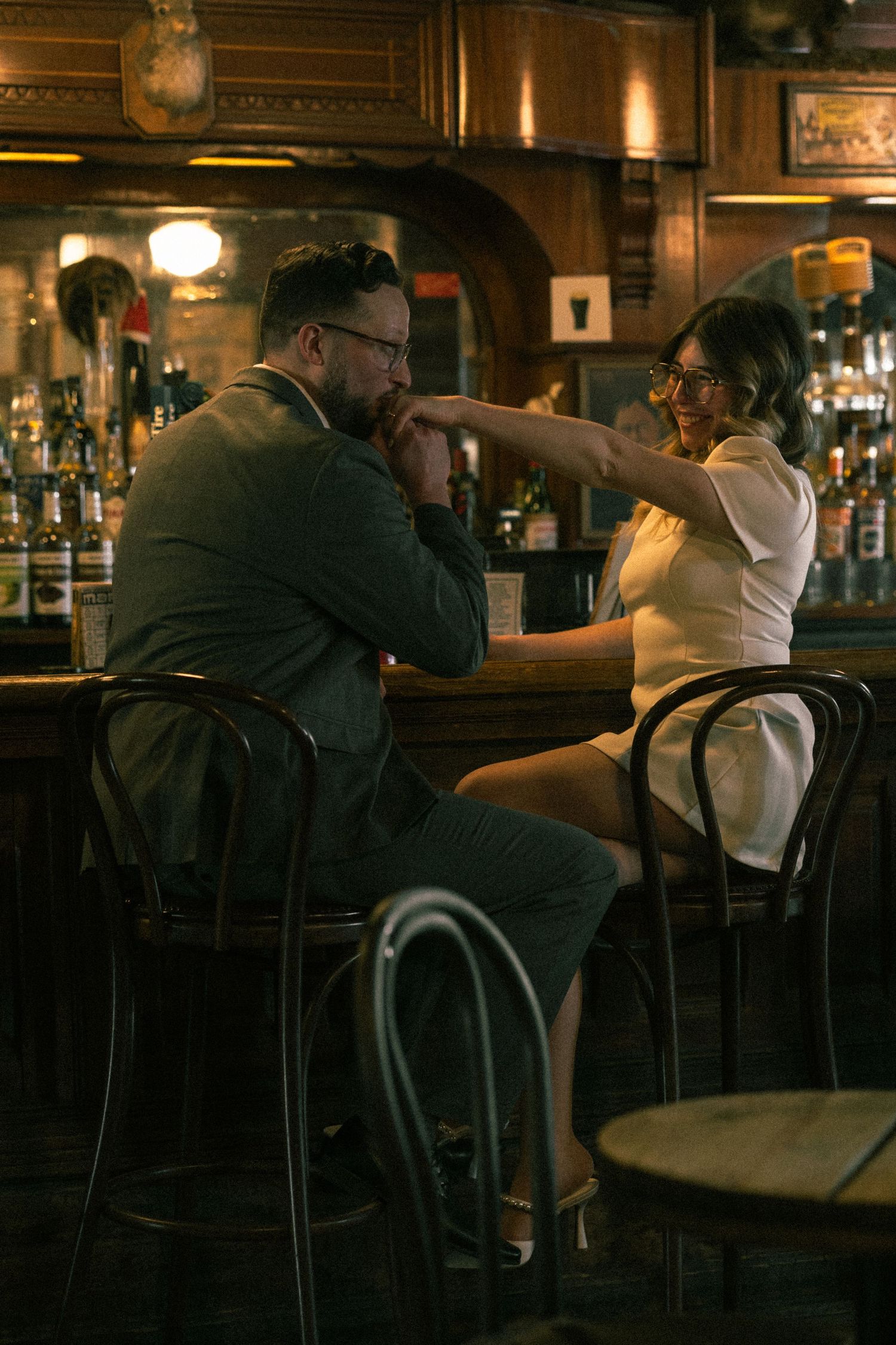 Couple sharing romantic moment at vintage bar, woman extending hand to man seated on barstool in warm amber light.