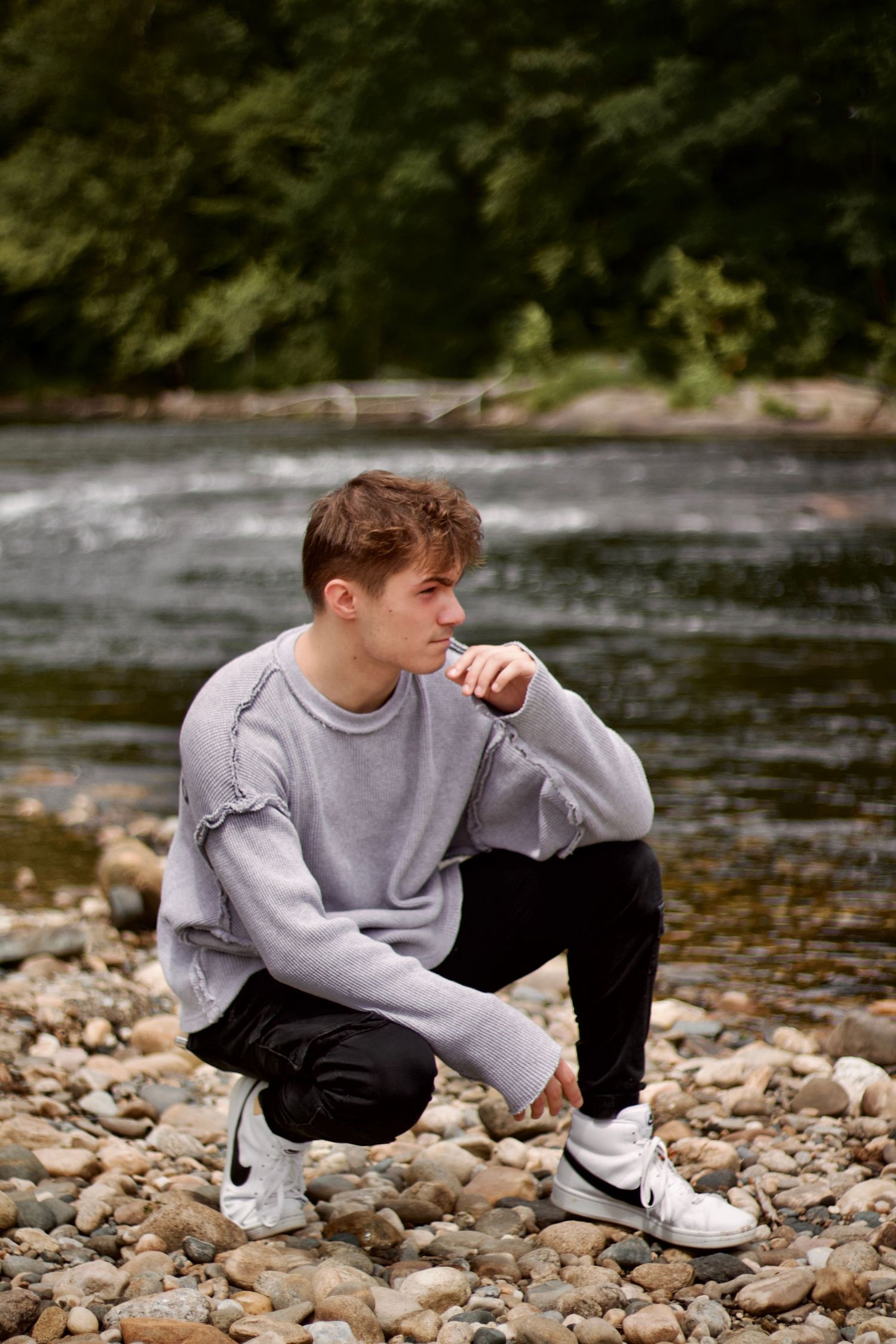 Westfield photographer photo of a Person crouching on rocky ground wearing gray sweatshirt and black pants with flowing river in background