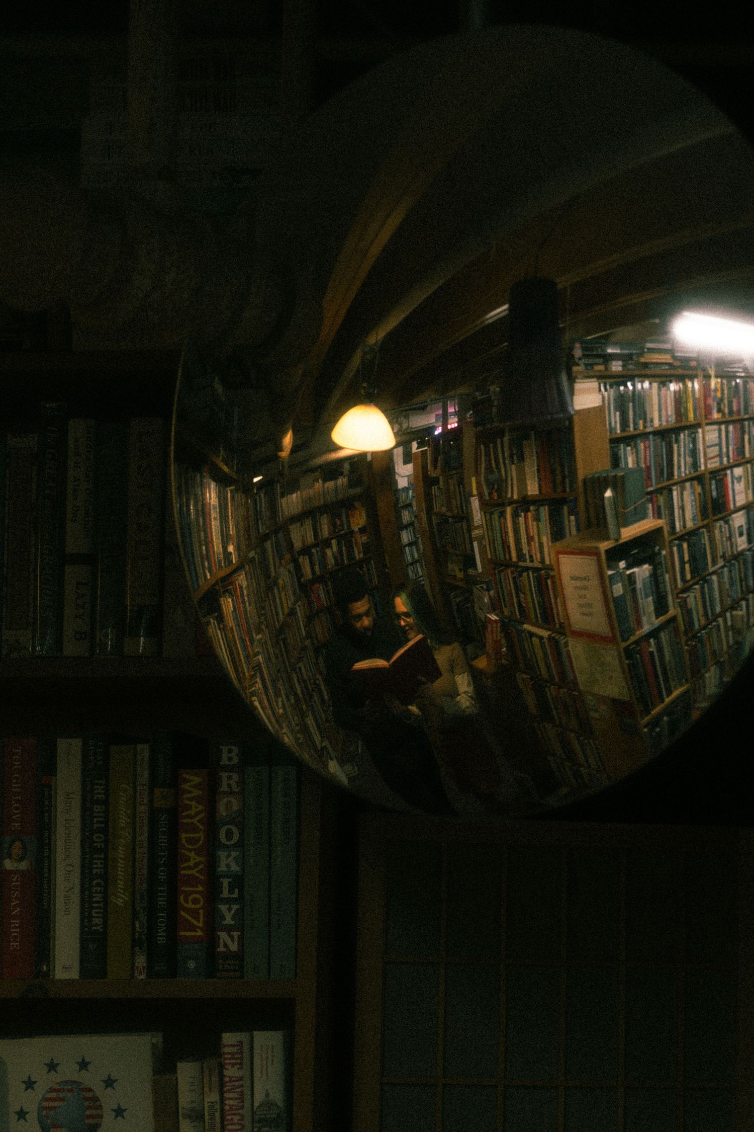 A convex security mirror reflects bookshelves and a reading area in a dimly lit bookstore or library.