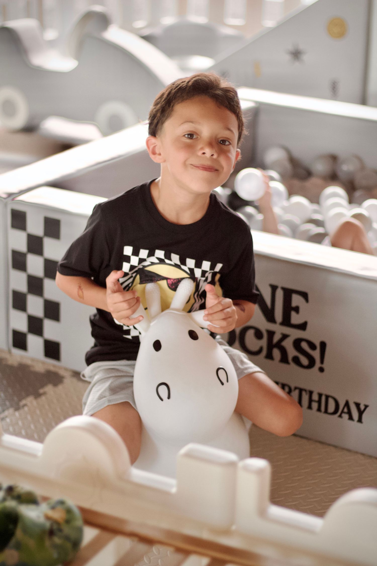 Young child in black checkered shirt sitting and playing with white balloon at birthday celebration.