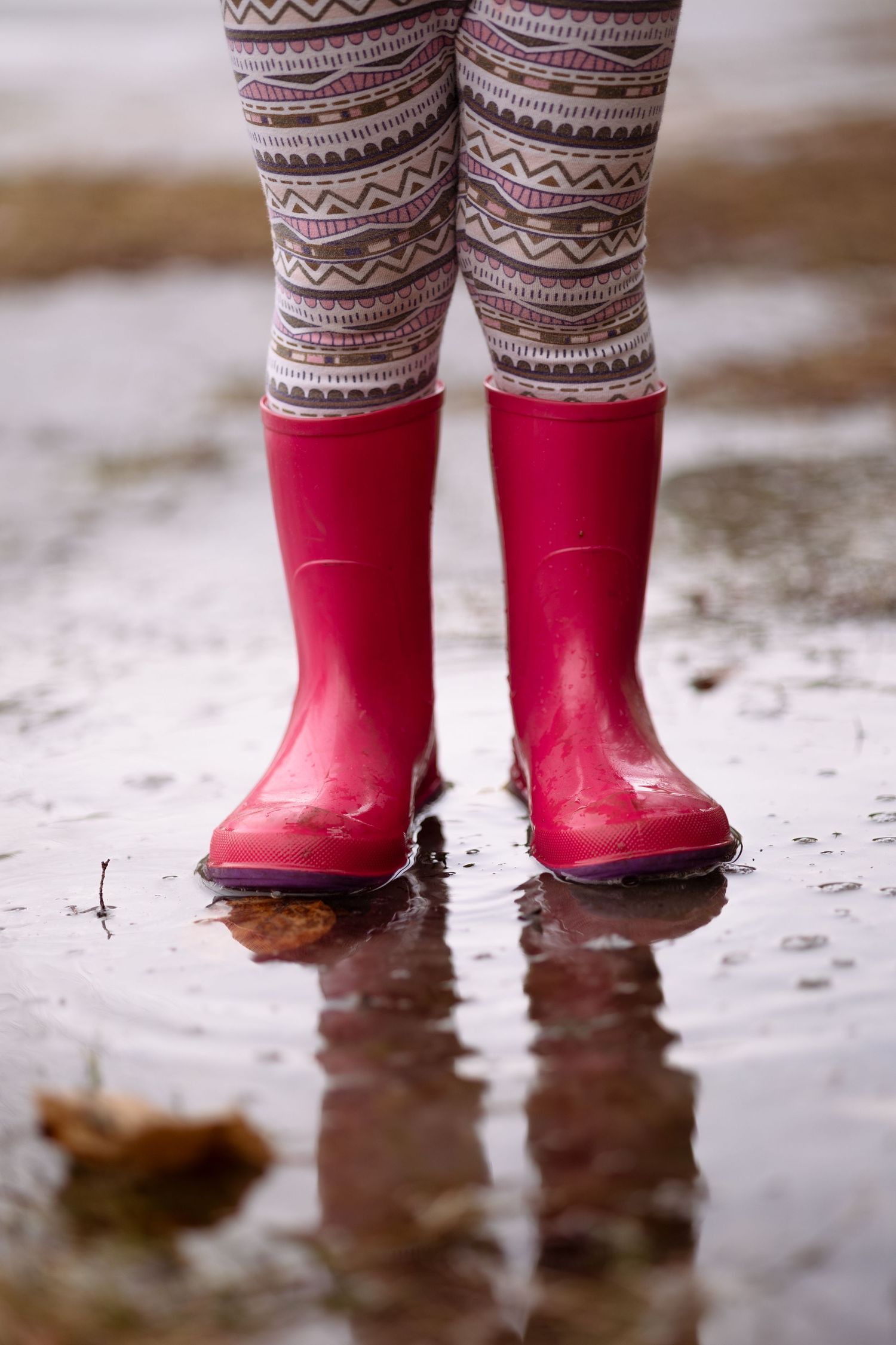 A pair of bright red rain boots with patterned leggings stands in a shallow puddle on a rainy day.