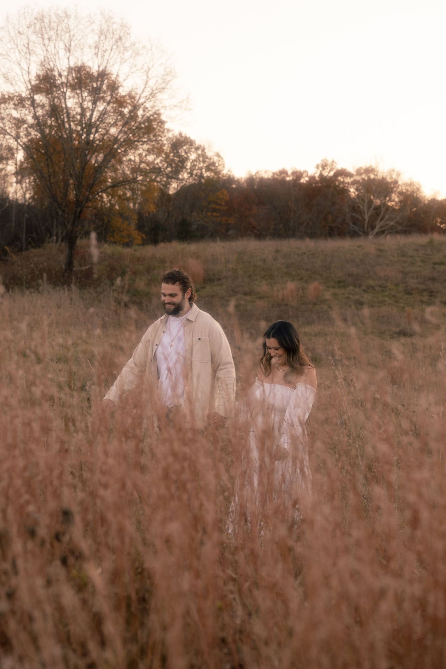 A couple stands together in a golden autumn field at sunset surrounded by tall dried grass and bare trees.