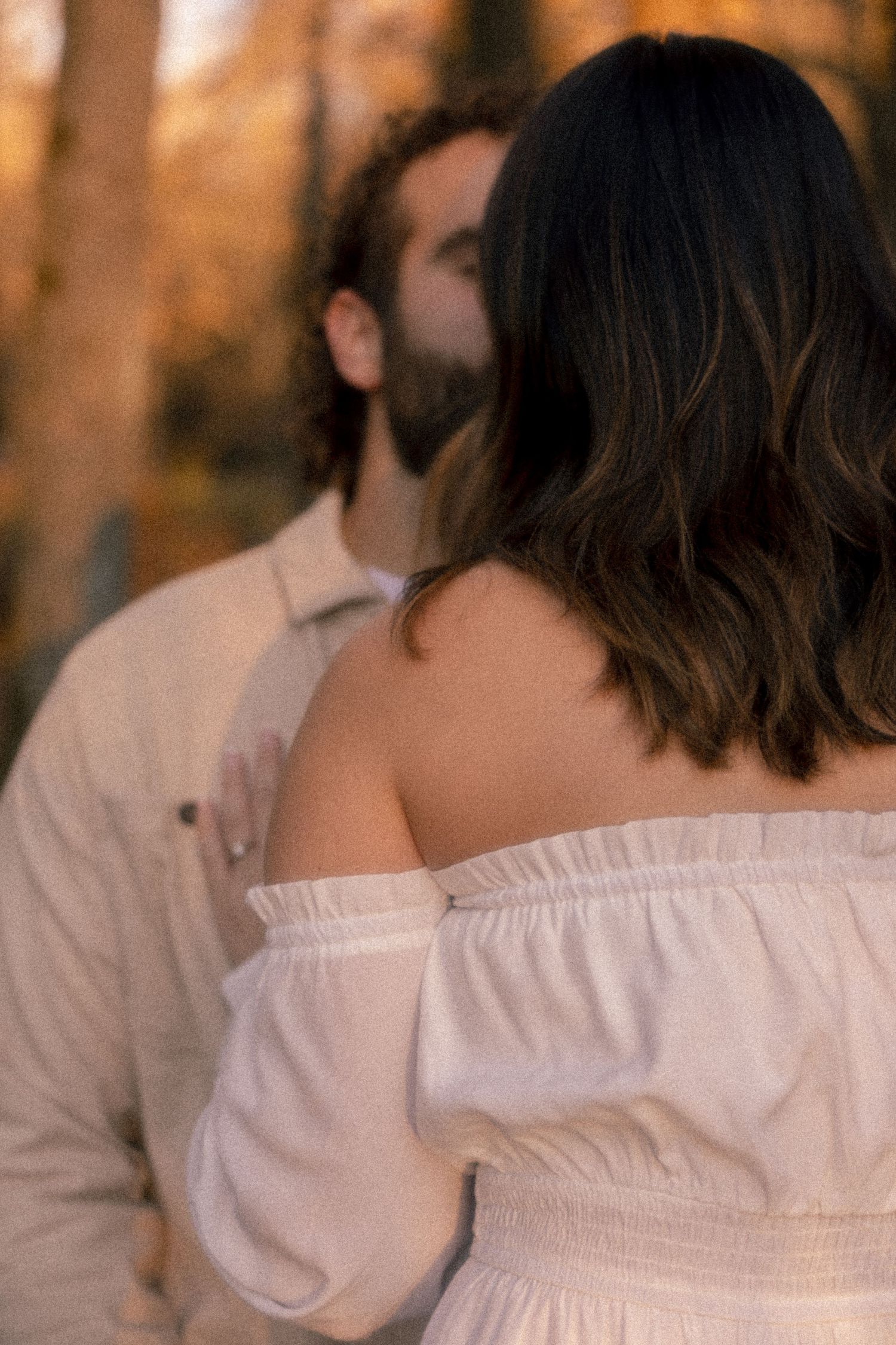 A couple in white clothing stands close together during an autumn photoshoot with warm orange tones in the background.