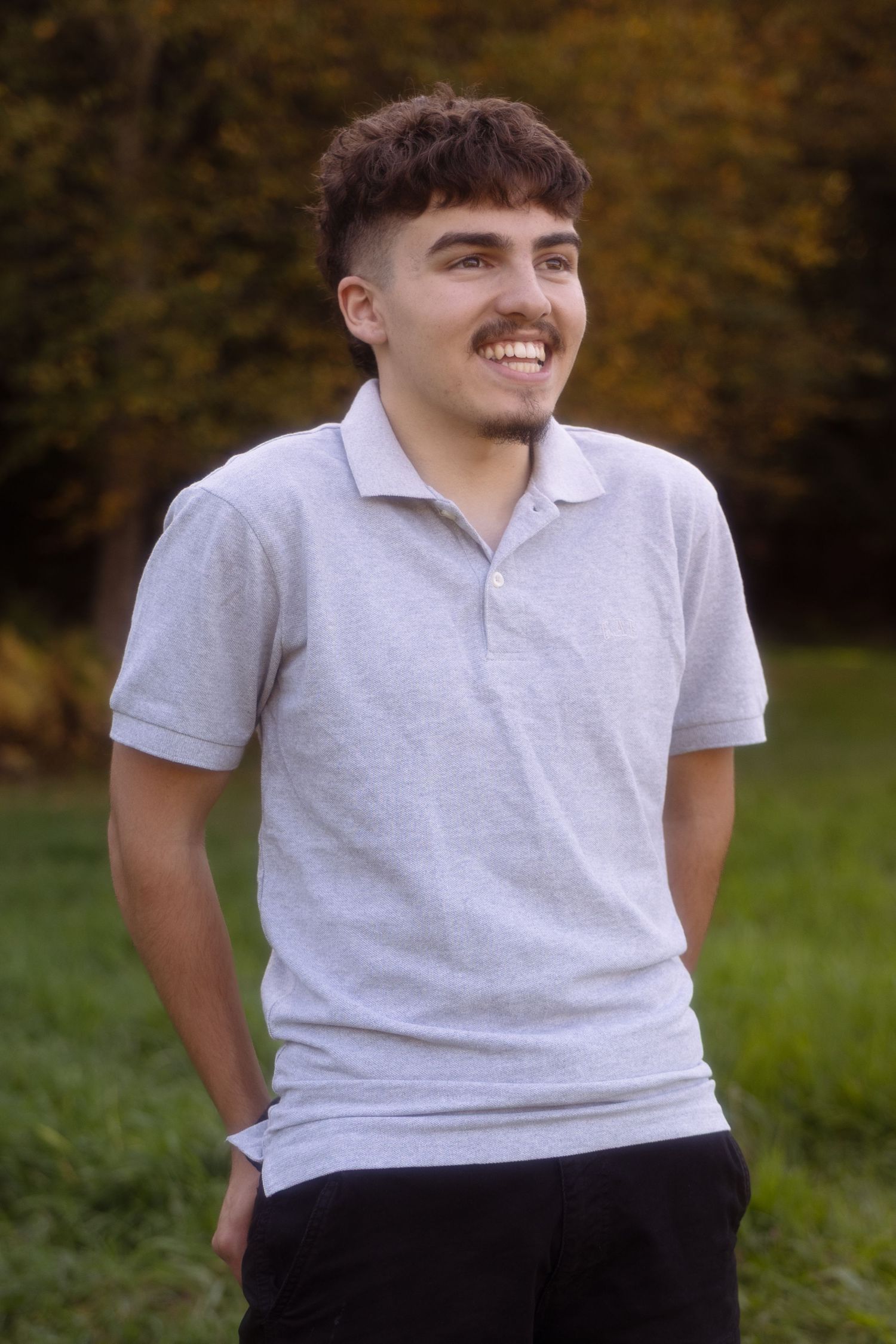 A person in a white polo shirt and dark pants smiles while standing outdoors against an autumn background.