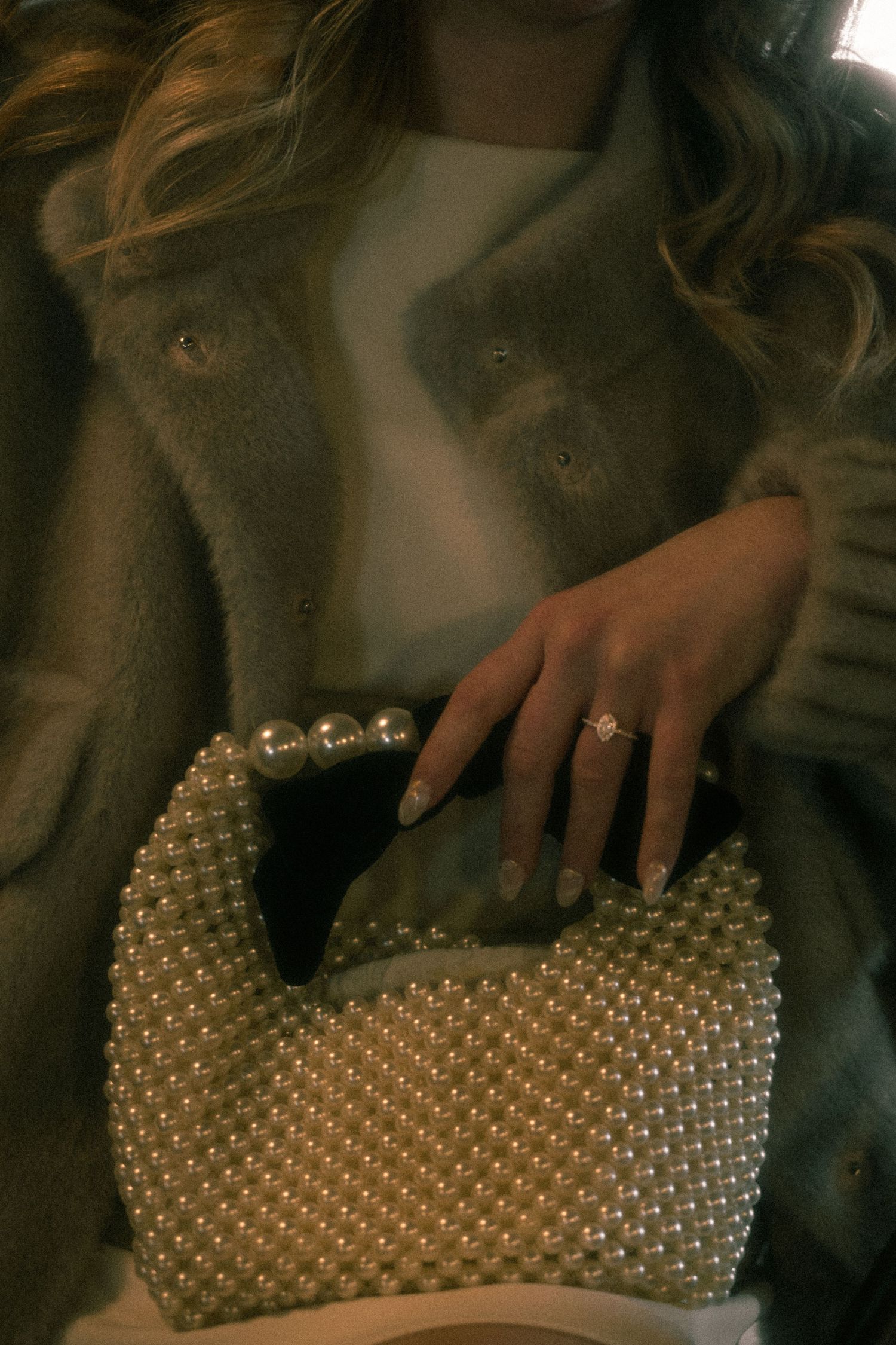Woman holding a pearl-beaded clutch bag, wearing a fuzzy coat and diamond ring with French manicure nails.